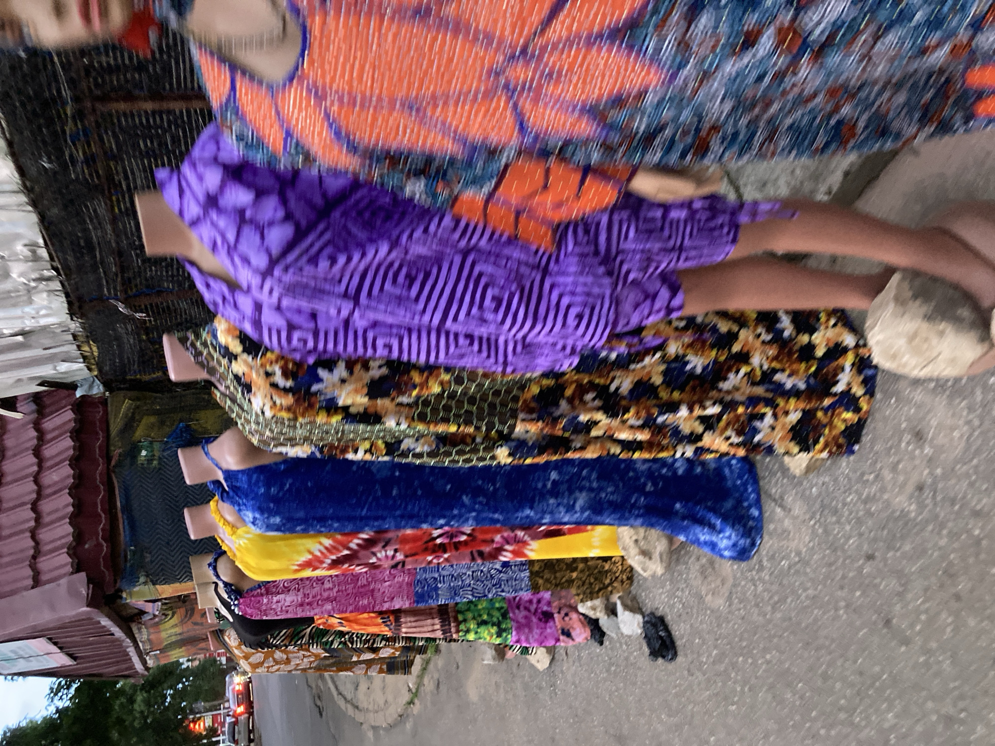 A street-side market stall displays colorful dresses on mannequins in Jos, Nigeria. The stall, covered by a reddish-brown corrugated metal roof, is set up along a paved street. Various patterned dresses, primarily in vibrant blues, yellows, purples, and oranges, hang on mannequins lined up against the stall. Some dresses feature floral prints, while others have geometric designs. The ground in front of the stall is a gray asphalt road, with scattered rocks being used to hold down the dresses. In the background, a few cars are visible on the road, and greenery can be seen further in the distance, suggesting an urban environment. The lighting indicates it is likely late afternoon or early evening.