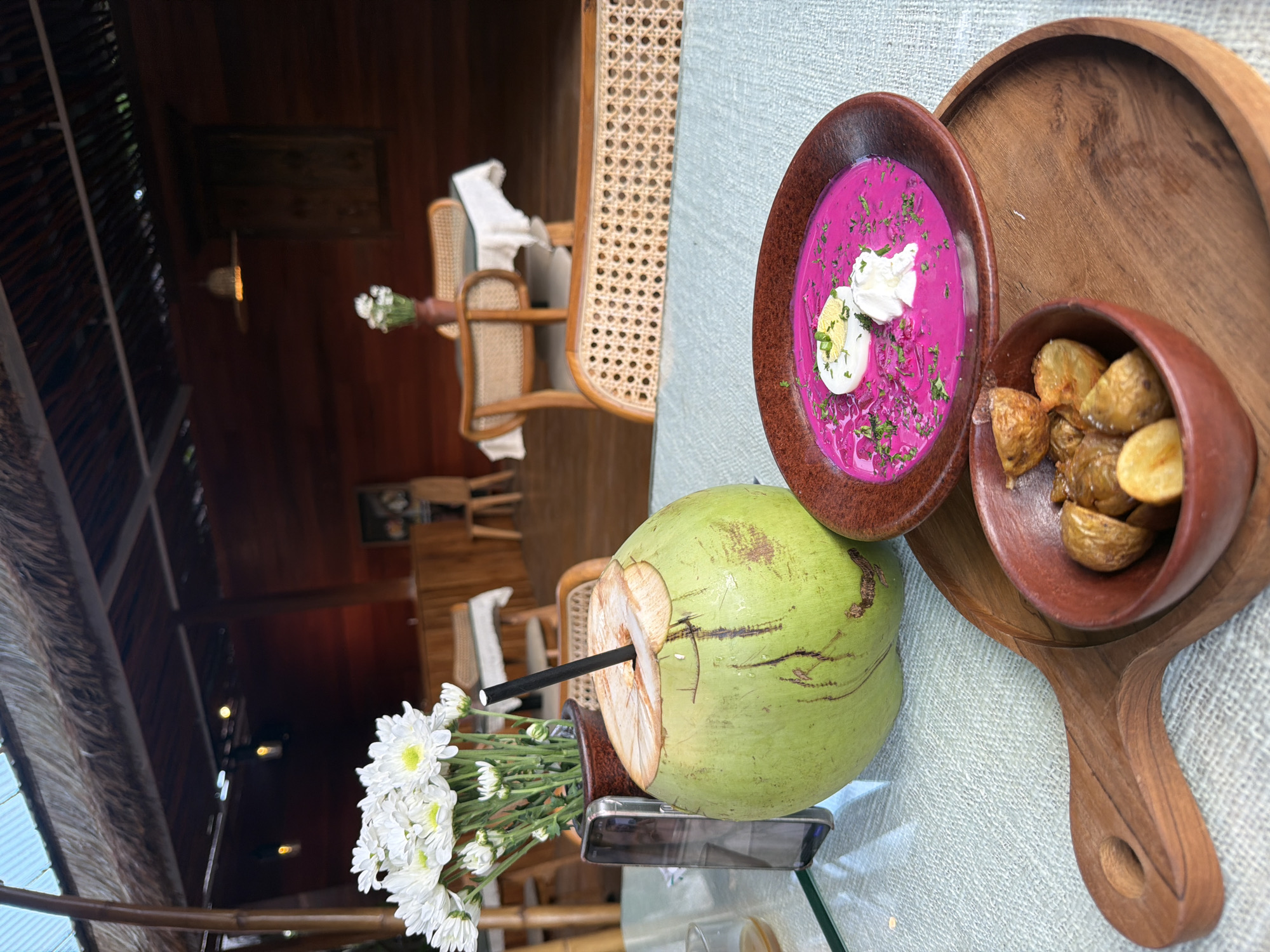 The image captures a meal setting, likely in a restaurant or café, in Petulu, Indonesia. A round wooden serving tray features a bowl of cold borscht with half of a hardboiled egg and herbs on top, along with a small bowl of roasted potatoes. Next to the tray is a young coconut with a straw, sitting on a table with a small vase of white daisies and a cell phone. The background showcases a rustic interior with wooden walls, furniture, and thatched roofing, indicative of the local architectural style. Rattan chairs are visible near tables set for dining, suggesting a relaxed ambiance.