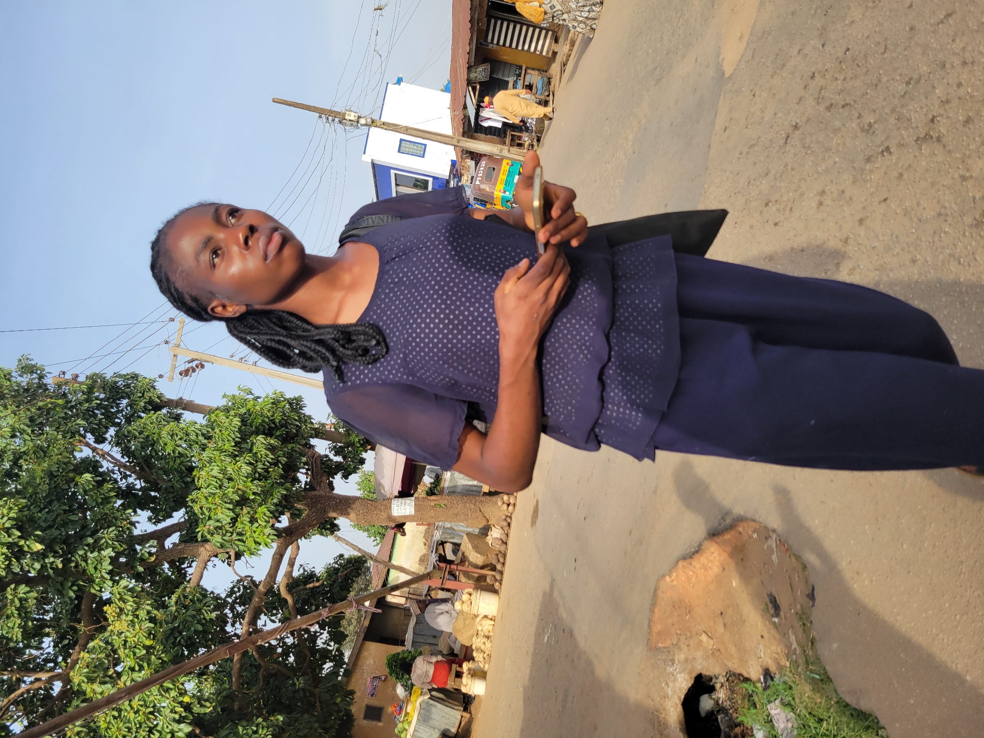 A young woman with braided hair stands on a paved street in Jos, Nigeria, holding a smartphone. She is wearing a dark blue, polka-dotted top and matching pants. She appears to be looking upwards, away from the camera. Behind her, there is a large leafy tree, utility poles with wires, and various buildings and shops along the street. In the foreground, there's a partially broken concrete structure on the roadside. A market is situated in the background under a corrugated iron roof. A tricycle painted green, yellow and white is also in the background.