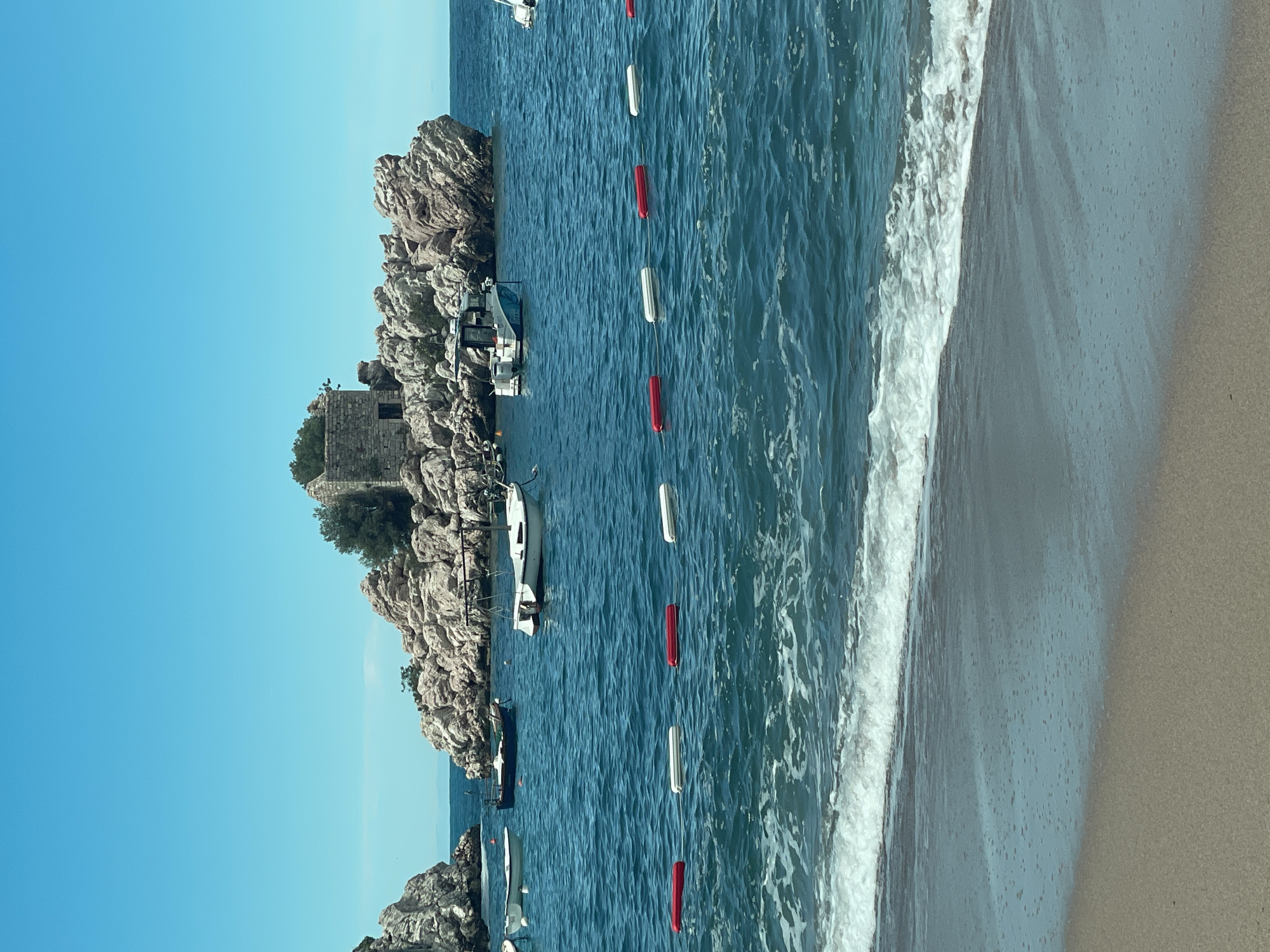 The image shows a coastal scene with a rocky island and a beach. The island features a stone structure, possibly a tower, amidst the rocks and sparse vegetation. Several boats are anchored near the island. A line of red and white buoys stretches across the water, potentially marking a boundary. The beach is sandy with waves gently breaking along the shoreline. The sky is clear and blue. The location is in the city of Unknown, Unknown.