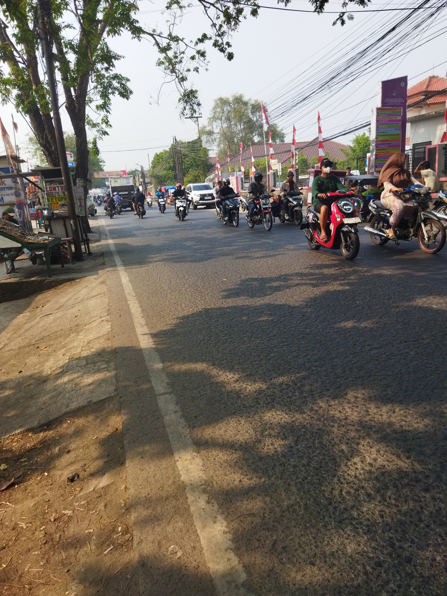The image captures a street scene in Sepatan, Indonesia, under a bright sky. The road is populated with various vehicles, primarily motorcycles, suggesting a typical day of traffic. Several individuals are seen riding motorcycles, some wearing helmets and masks, indicating health precautions or a preference for protective gear. One person in the foreground is riding a pink scooter. To the left of the road, there are street vendors and shops with signage in Indonesian, hinting at local commerce. The presence of Indonesian flags and what appears to be a building with institutional signage suggests a public or governmental area. The environment is characteristic of a bustling urban or semi-urban setting in Indonesia, with trees lining parts of the street and electrical wires overhead. The sunlight casts shadows on the road, indicating it is daytime.