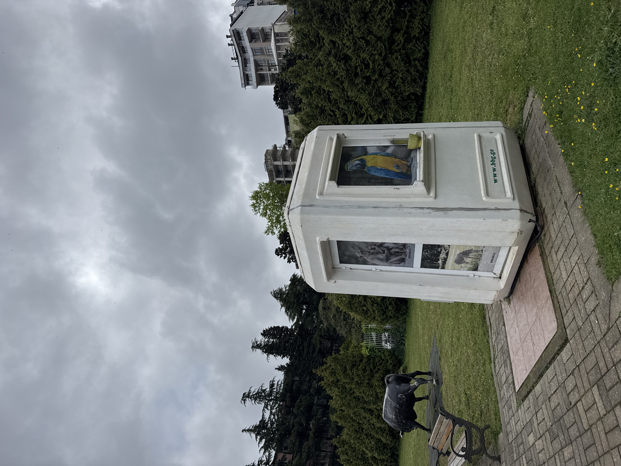 Captured from a low-angle perspective under an overcast sky, the image presents a kiosk-like structure standing on a small tiled platform adjacent to a brick pathway. The kiosk is adorned with images, including one of a blue and yellow parrot, visible through its window. A metal sculpture of an animal, possibly a bull, stands near a bench placed in front of the kiosk. The background features trees and the upper floors of a building. The kiosk bears the inscription "www.bbg.ge." The scene is located in the city of Unknown, Unknown.