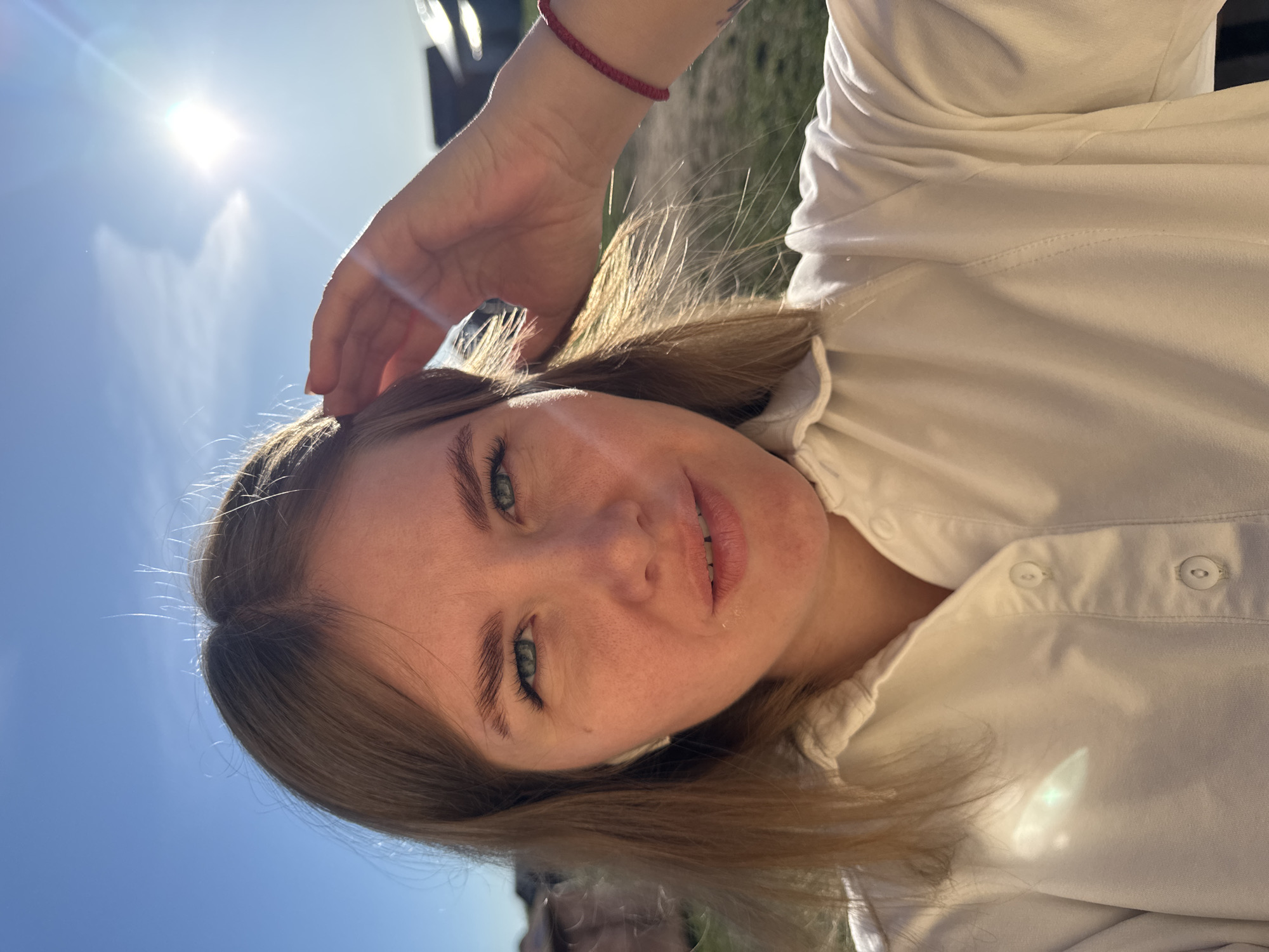 A young woman with light brown hair and green eyes poses for a selfie in Ферма, Russia. She wears a white button-down shirt and a red bracelet on her wrist. Her hand rests on her head. The bright sun and blue sky are visible in the background. The setting appears to be outdoors, possibly in a park or grassy area.