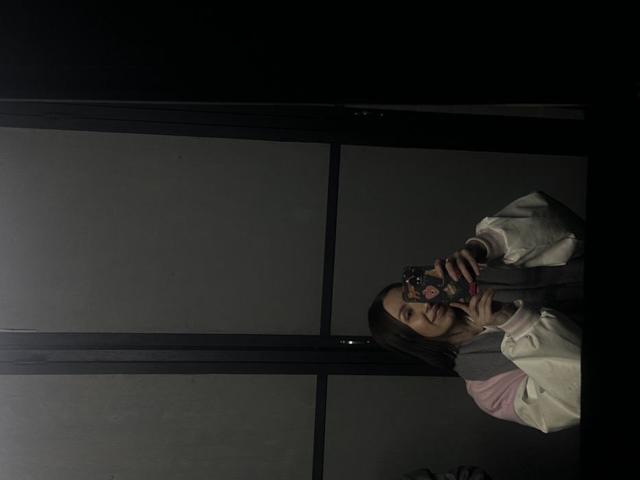 A female individual, appearing to be in her twenties or thirties, is captured in a mirror reflection, holding a smartphone to take a photograph. She is positioned centrally in the lower half of the frame, smiling faintly. Her hair is dark brown and straight, reaching just below her shoulders. She wears a light-colored, possibly white or cream-colored, jacket with distinct pink upper arm or shoulder sections, and a grey scarf wrapped around her neck. The smartphone has a dark-colored case adorned with multiple small, colorful designs, including what appears to be a heart and animal prints. The background visible in the mirror behind the subject consists of flat, dark grey panels segmented by dark, slender vertical and horizontal metallic or wooden frames, suggesting an interior wall, partition, or door system. The overall lighting of the scene is dim. The specific location within the city of Izhevsk, Russia, is not identifiable from the visual information provided in the image.