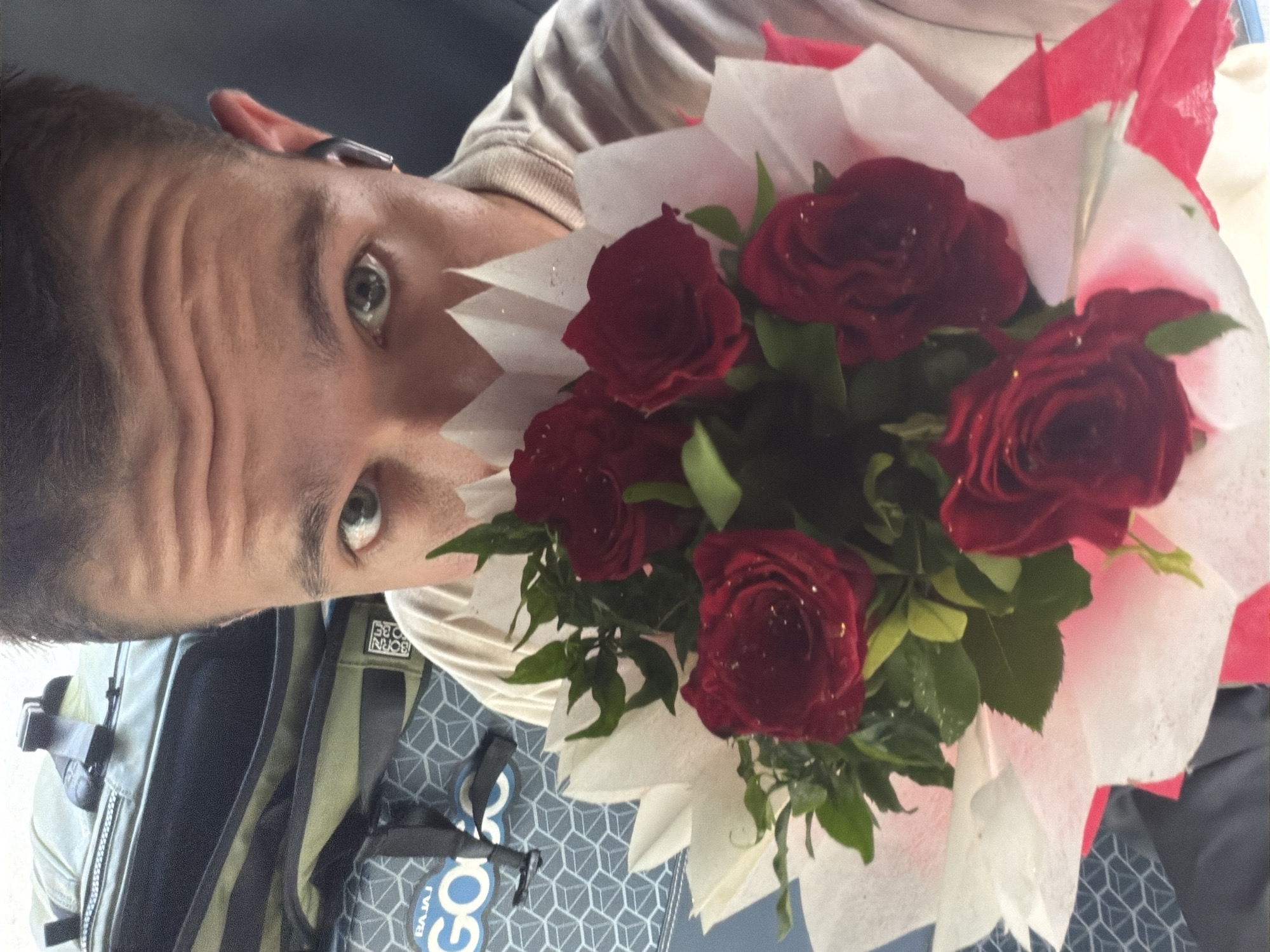 A close-up, selfie-style shot shows a young man holding a bouquet of red roses in front of his face. He is wearing a light-colored sweatshirt and has a subtle smile visible around his eyes. A dark-colored earbud is in his right ear. In the background, a green duffel bag with the "BAJAJ GO" logo is visible. The man's face is partially obscured by the bouquet, which is wrapped in white and pink paper. The lighting is natural, suggesting an outdoor or well-lit indoor environment. The presence of the duffel bag and the man's attire hint at travel. The image does not provide any specific landmarks or details to pinpoint a location within Kanpur, India.