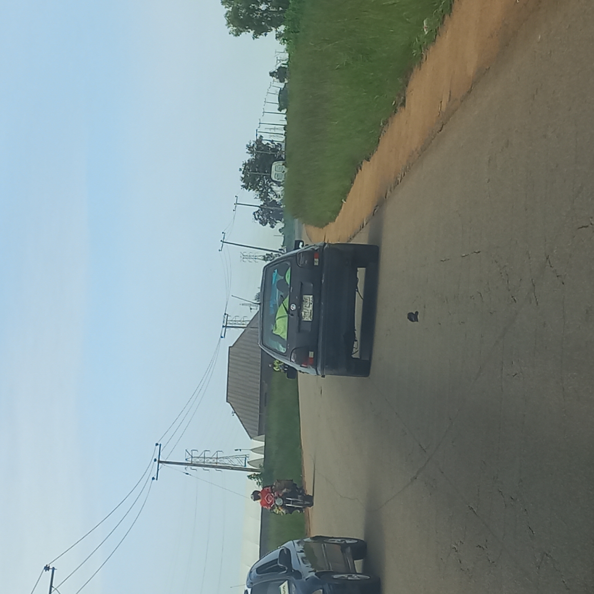 The image depicts a paved road in the city of Kassa Market, Nigeria. The road is flanked by green grass and foliage on the right side, and the left side shows a dark-colored car. In the distance, there are utility poles with power lines stretching across the sky, which is a pale blue with some clouds. A black Volkswagen Golf is visible in the foreground, parked or moving slowly on the road. A motorcycle with a rider wearing an orange shirt is also present on the road. There are buildings in the background, including a structure with a corrugated roof. The road surface shows signs of wear and tear, with visible cracks.