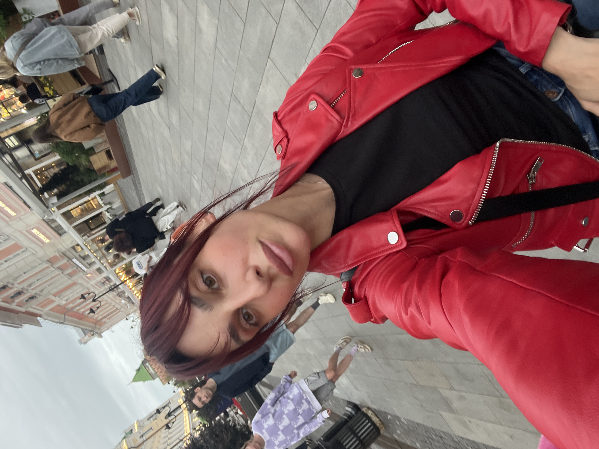 A woman is taking a selfie on a city street in Nizhny Novgorod, Russia. She is wearing a red leather jacket, a black t-shirt, and blue jeans. In the background, other people are walking on the sidewalk. The street is paved with stone tiles, and there are buildings on either side. The sky is overcast.