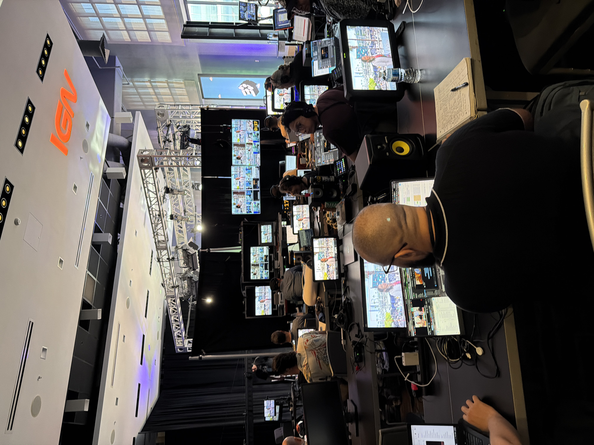 The image depicts a scene inside a television broadcast studio or control room. Several individuals are seated at desks, many wearing headphones, suggesting they are working on a live production. Multiple monitors are visible, displaying video feeds, graphics, and what appear to be production schedules or control interfaces. The overall environment is professional and technical, with a focus on video production equipment. The IGN logo is prominently displayed on the ceiling, indicating the company's presence. The setting is likely a studio in San Diego, United States of America, a location known for its media and entertainment industry presence.