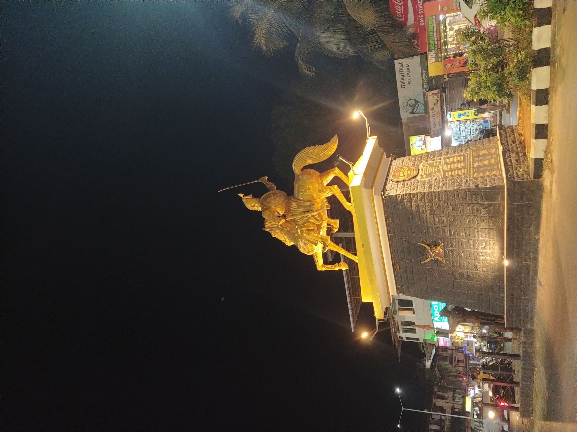 A golden statue of a man on horseback dominates the scene, likely located in Oxel, India. The statue is mounted on a raised, yellow-and-gray stone base, which also features inscribed plaques. The background includes a mix of urban elements: a street with visible sidewalks, lit by streetlights, alongside buildings and commercial establishments featuring signs such as "Milky Mist Ice Cream," "Coca-Cola Café Street," and "Apol." Palm trees are seen above some of the buildings. The sky is dark, indicating it's nighttime.