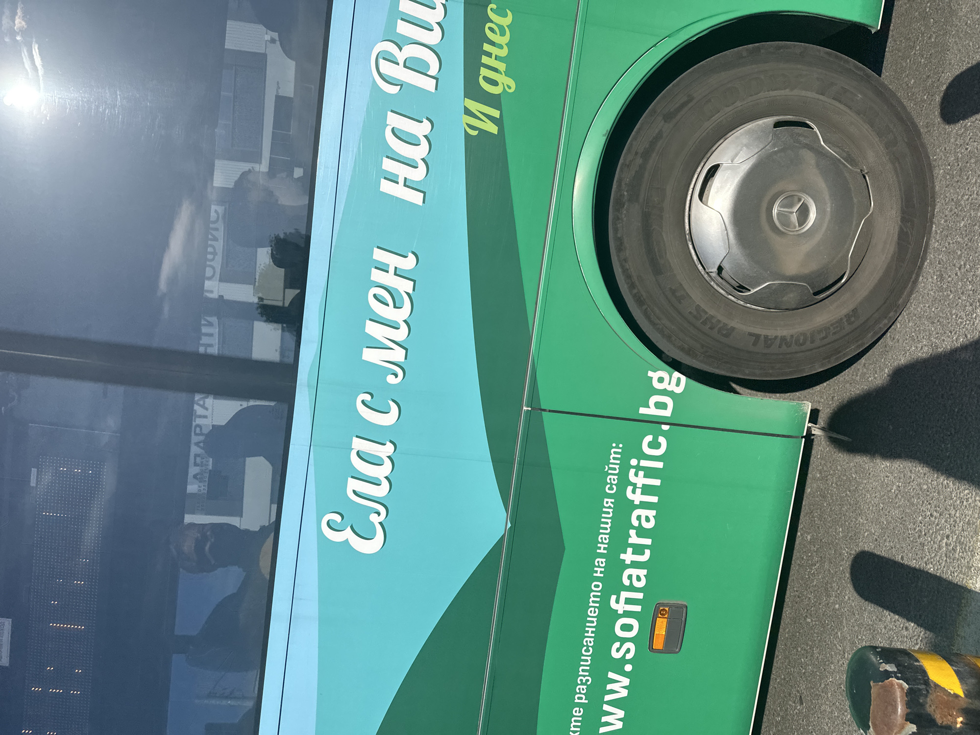 This image shows a portion of a green bus in Sofia, Bulgaria. A passenger is visible in the bus's window. The bus has text in Cyrillic that reads "Ела с мен на Ви...И днес," which translates to "Come with me on Vi...And today." Below that, in smaller text, it says "www.sofiatraffic.bg". The bus's tire and wheel hub are visible, showing a Mercedes-Benz logo. The word "adaptar" is faintly visible in the reflection on the bus window.