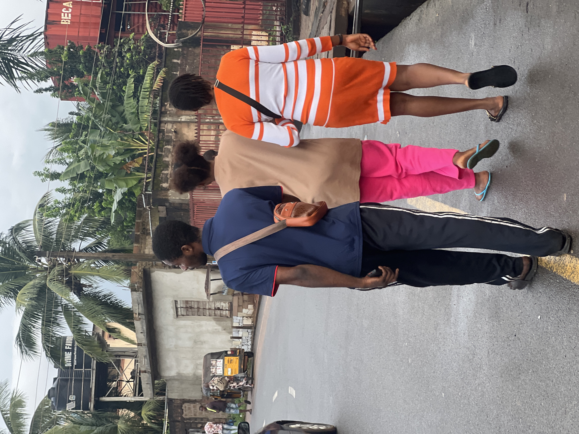 In Nkpor, Nigeria, three individuals walk along a wet asphalt road. A man in a blue t-shirt and black track pants is in the foreground, carrying a brown shoulder bag. Behind him, two women walk side-by-side. The woman on the left wears a light brown long-sleeved shirt and bright pink pants, while the woman on the right is dressed in a long-sleeved orange and white striped dress. In the background, a yellow auto-rickshaw is parked, and buildings with water tanks are visible. Palm trees and lush vegetation are scattered throughout the scene, suggesting a tropical urban environment. The sky is overcast.