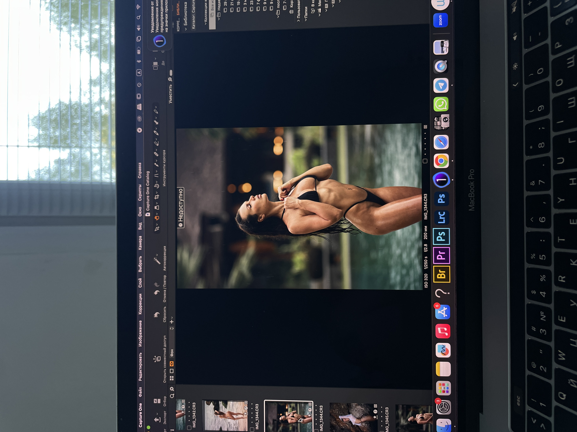 The image shows a MacBook Pro screen displaying the Capture One software with a photograph of a woman in a bikini. The woman is lying on her side in what appears to be a shallow body of water with a blurred background of greenery and lights. The Capture One interface includes menus (File, Edit, Image, Corrections, Layer, Select, Camera, View, Window, Scripts, Help) and toolbars. The bottom of the screen shows several thumbnail images of other photos in the same folder. The MacBook Pro is surrounded by the user's surrounding environment, including blinds covering a window and the keyboard. There is no information in the image indicating the location is in Kazan, Russia.