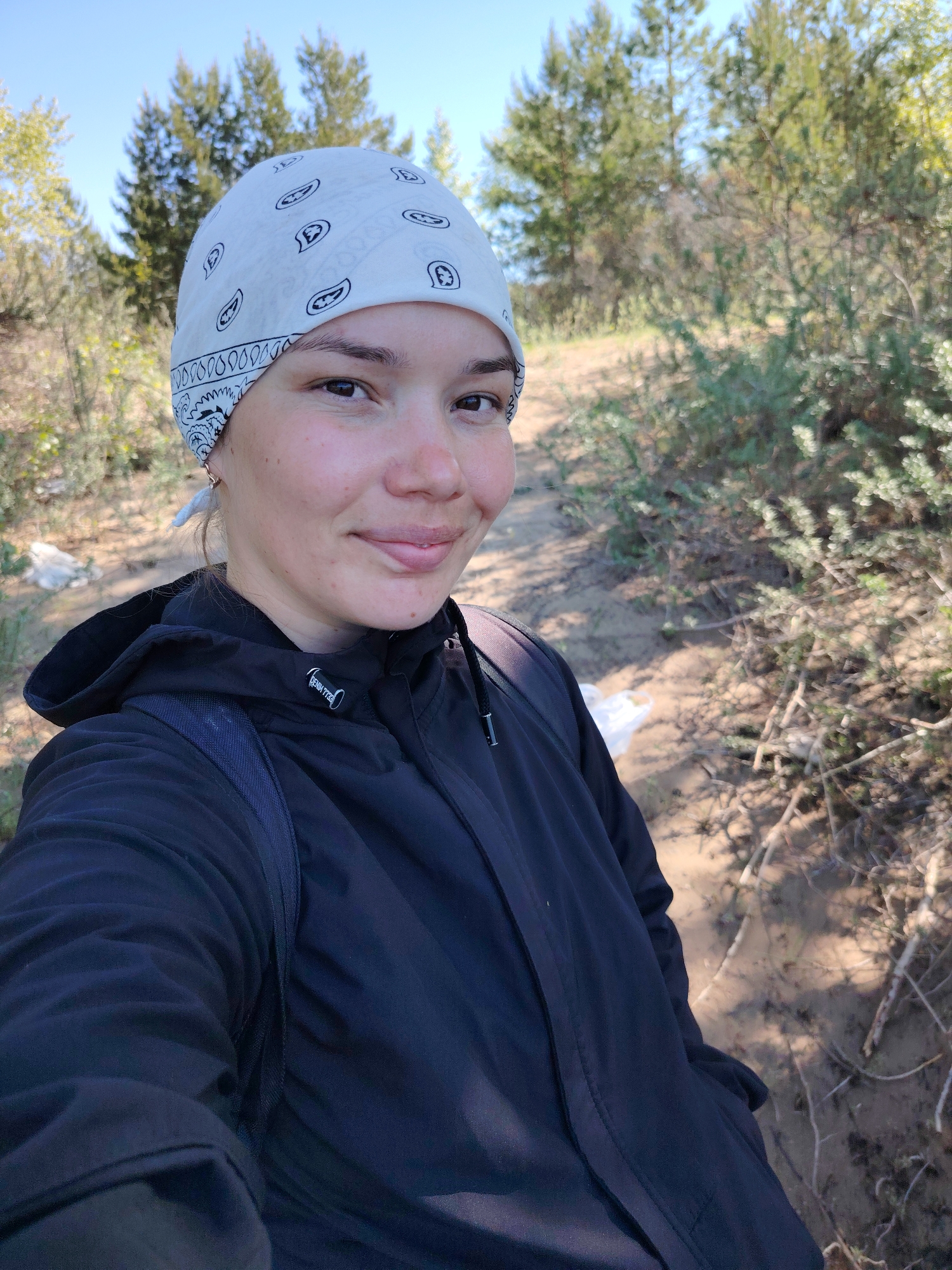 A young adult female takes a selfie outdoors, wearing a white bandana with a paisley print and a dark blue hooded jacket. She is smiling softly. The background features trees and sandy terrain with some shrubbery. The location appears to be a natural area, potentially a park or a wooded area. Information about the precise location being in the city of Chaikovsky, Russia, is not discernible from the visual data.
