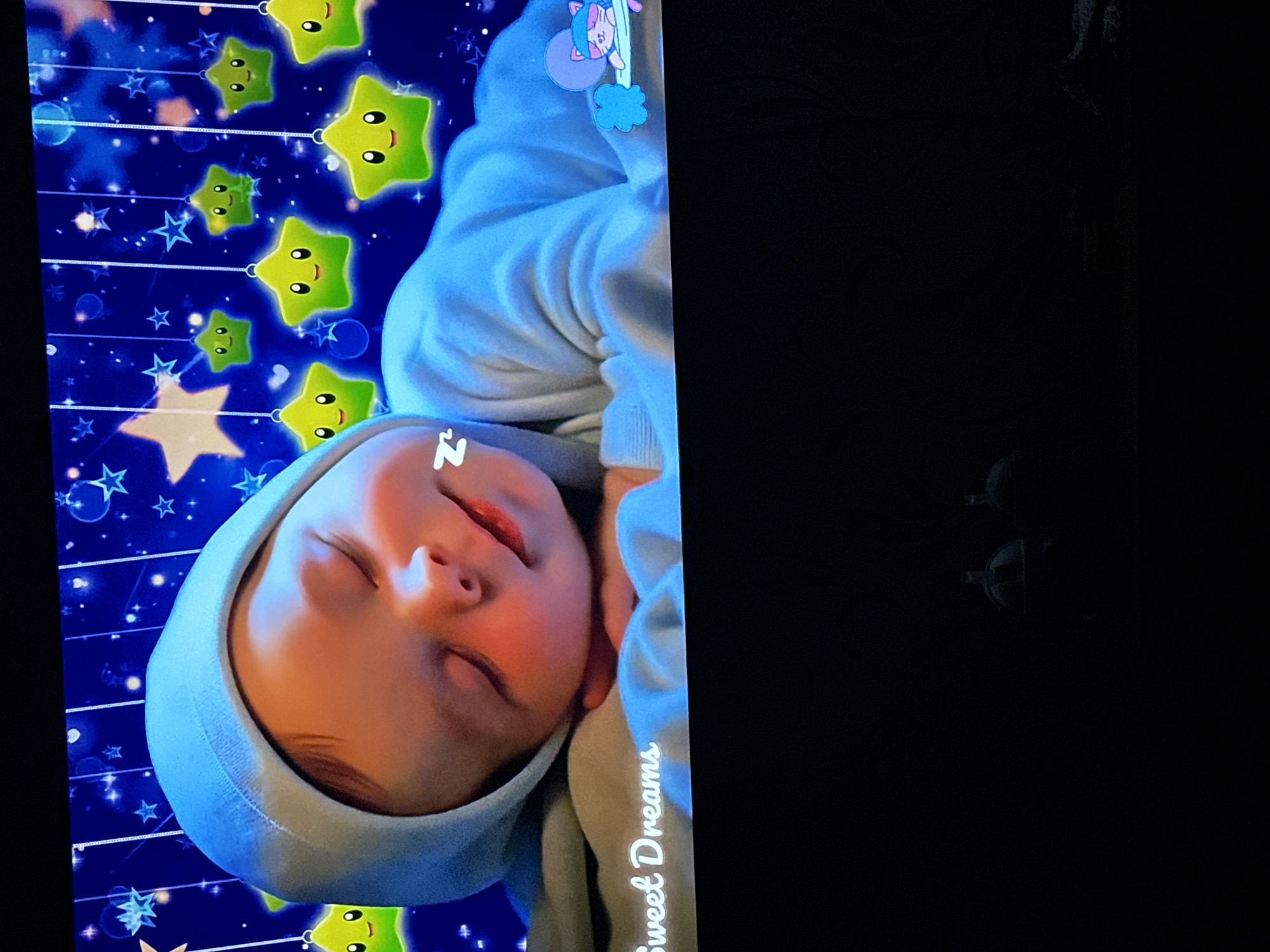 A television screen displays a sleep-themed animation. In the foreground, a close-up shot shows a sleeping baby wearing a light blue hooded onesie. The baby is lying on its side, with its head resting on its arms. A stylized "Zzz" symbol hovers near the baby's head. The background features a dark blue, starry night sky with several cartoonish, smiling yellow stars hanging from strings. Towards the bottom right, a small animated cat wearing a pink sleeping mask is depicted lying on a crescent moon. The words "Sweet Dreams" are written in white script across the bottom left of the screen. The television itself is dark and appears to be in a room with very low ambient light. No specific location in the city of Comrat, Moldova can be determined from this image.