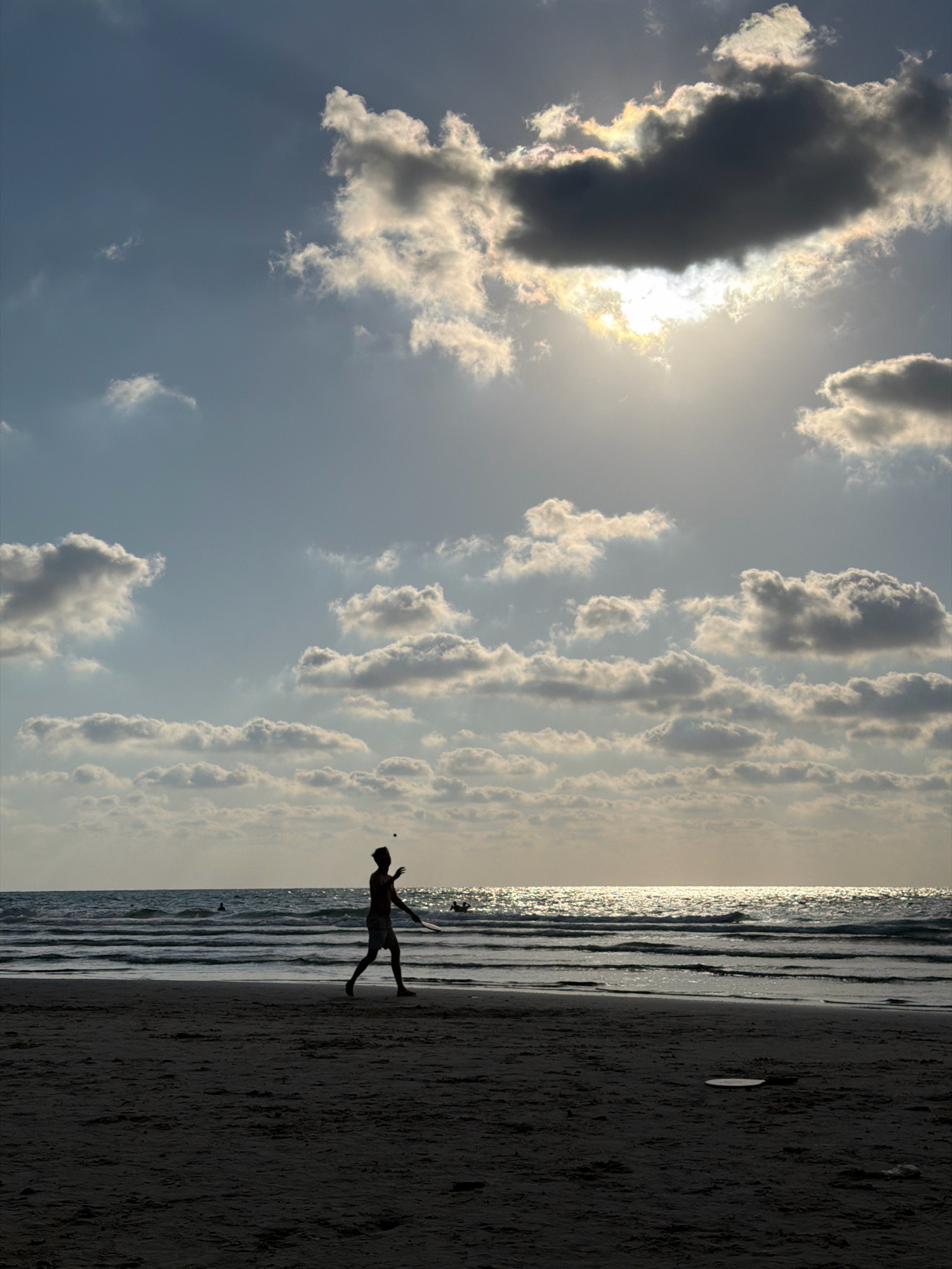 The image depicts a sunset scene at the Hadera beach in Israel. The sky is filled with dramatic clouds, with the sun partially obscured by a large, dark cloud, casting beams of light and creating a luminous effect. Below the sky, the ocean reflects the sunlight, creating a shimmering effect on the waves. On the sandy beach in the foreground, a silhouetted figure of a person is captured mid-action, playing with a racket and a ball. The person is dressed in shorts and appears to be in motion, preparing to hit the ball. Further back, in the water, other indistinct figures can be seen, suggesting the presence of other beachgoers. The overall mood is serene, with the natural beauty of the sunset and the activity of the lone figure on the beach contributing to a tranquil atmosphere.