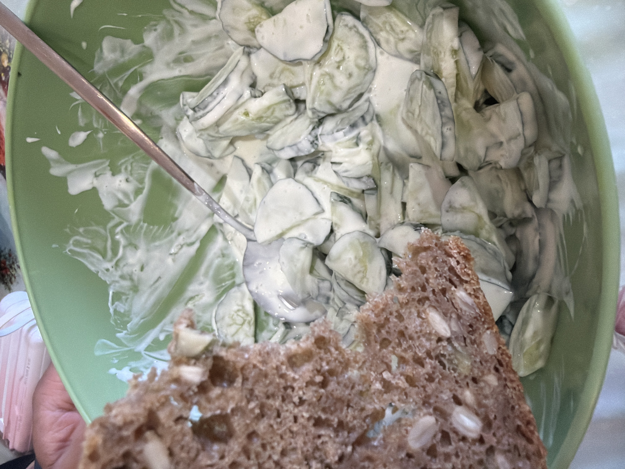The image shows a close-up of a light green bowl filled with cucumber salad coated in a white creamy dressing. A silver spoon is partially submerged in the salad. In the foreground, a slice of brown bread with visible seeds is positioned in front of the bowl, slightly obscuring the view of the salad. The context of the location being in Chaikovsky, Russia is not visually evident from the image.