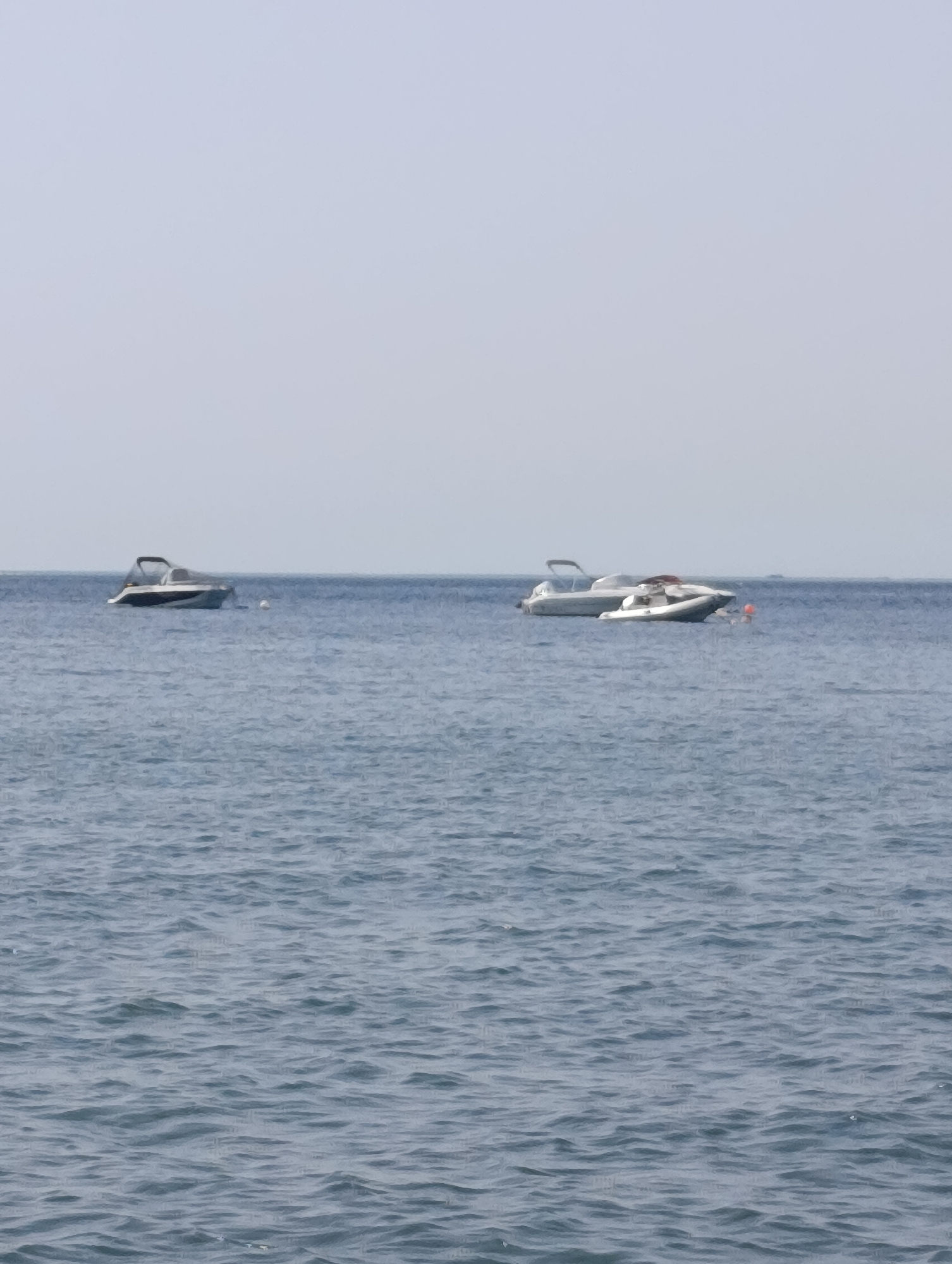The image shows a body of water, likely the Atlantic Ocean, with two motorboats at anchor. The water is choppy, indicating a light breeze or moderate sea state. The sky is overcast, with a uniform pale grey color. The boats are positioned at a distance from the viewer, with the left boat further away and the right boat closer. The boat on the right appears to be a larger cruiser, possibly with a cabin, while the boat on the left is smaller, possibly a speed boat. There is no indication of land or city in the background, suggesting the boats are offshore. The location is not specifically identifiable as Setúbal, Portugal, as the image lacks distinguishing landmarks or urban elements. The scene is static and peaceful, with no visible human activity.