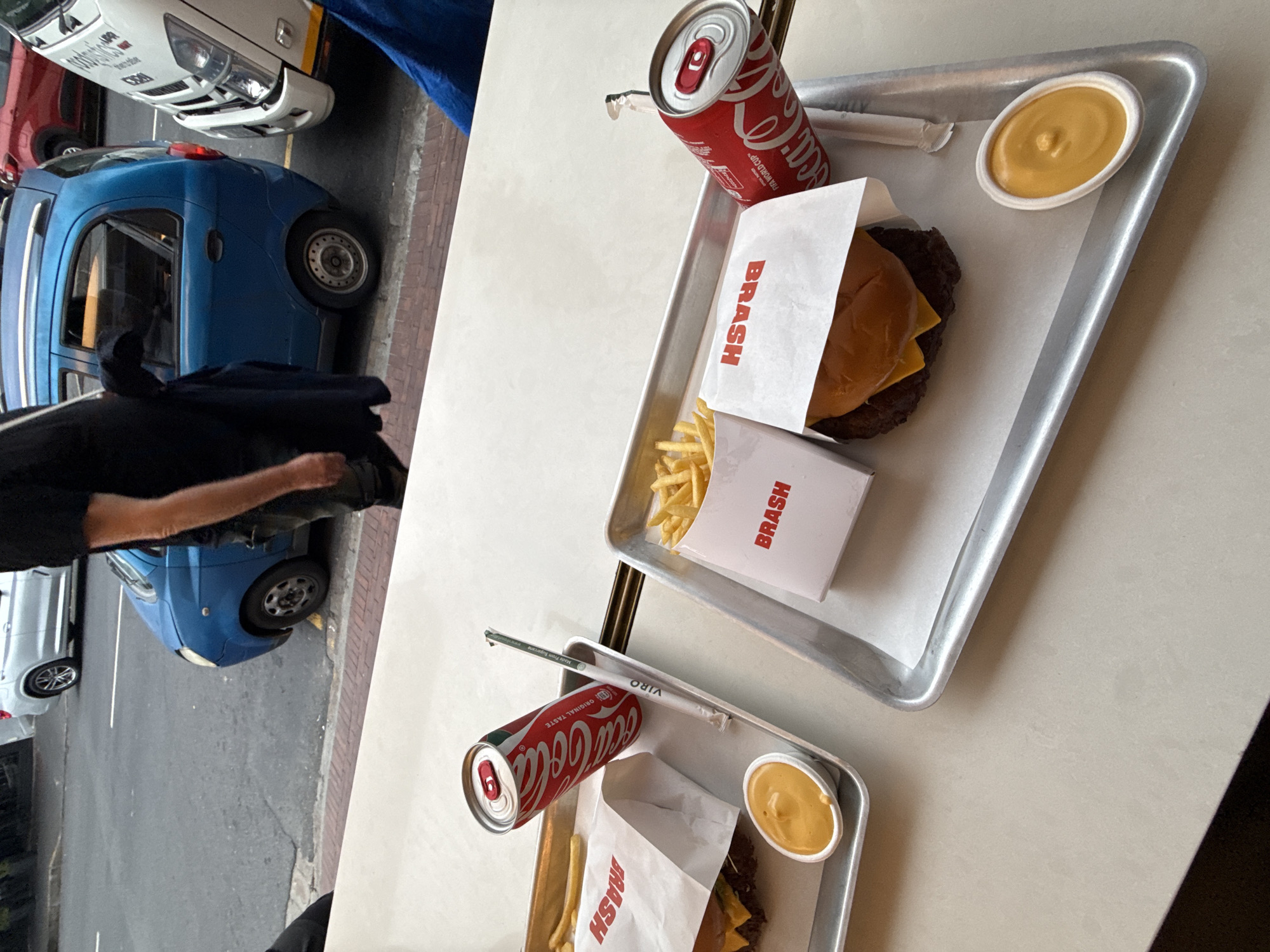 The image captures an aerial view of two trays on a light-colored table, each containing a fast-food meal. Both meals include a burger in a "BRASH" branded wrapper, a portion of fries also in a "BRASH" wrapper, a Coca-Cola can, and a small container of dipping sauce. The trays are simple, rectangular, and appear to be made of metal. In the background, a street scene is partially visible, featuring parked cars including a small blue hatchback, a red car, and a white vehicle, and a brick pavement. The presence of "BRASH" as a branding element suggests the location is Cape Town, South Africa, as this is a restaurant chain present in that city. A portion of a person's arm and black clothing is also visible, partially obscuring one of the vehicles. The image suggests a casual dining scenario, possibly outdoors, with a focus on the fast-food meals.