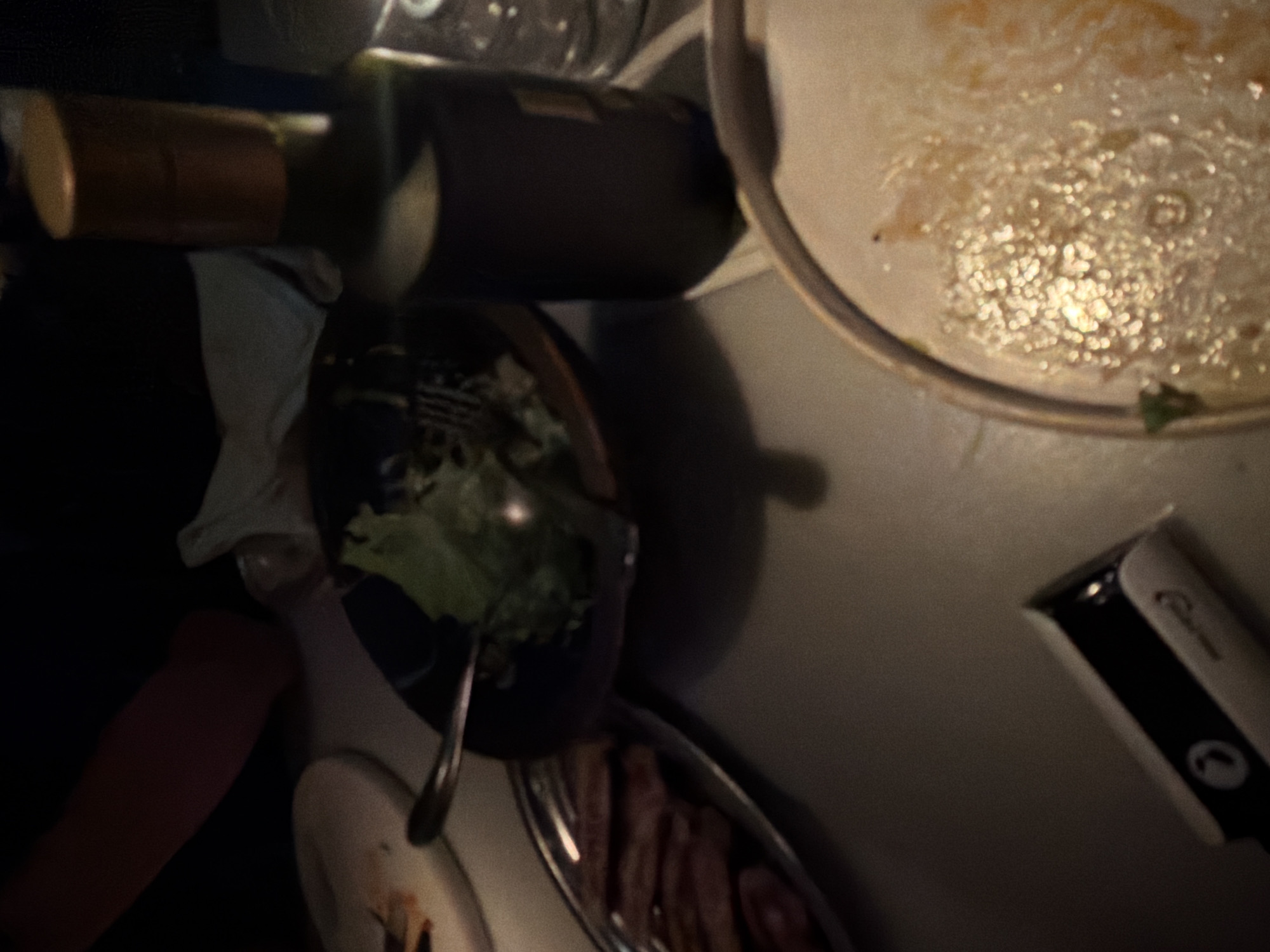 Here is a forensic description of the image:
The image appears to be a close-up shot, taken indoors. The angle is looking down at a table setting. A partially eaten plate of food is visible, along with a plate of what looks like meat slices. A bottle of wine lays beside the plates. A white object with black stripe is on the table edge, possibly an electric razor. The overall lighting is dim. There are no people directly visible in the scene, only traces of them through the remnants of the meal. The city and the location of the image cannot be determined from the available visual information.