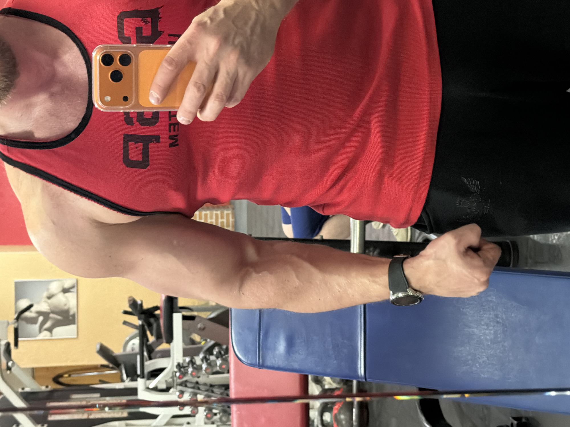 The image displays a close-up, mid-torso to upper-thigh view of a muscular male individual taking a selfie in a gym located in Saint Petersburg, Russia. The man wears a red sleeveless tank top with black trim, featuring a mirrored, partially obscured black logo on the chest that appears to read "GAP" backwards with "M.E.T." underneath. He is also wearing black athletic pants with a dark, stylized emblem on the left thigh. A silver-toned smartwatch with a black strap is visible on his left wrist. He holds an orange-cased smartphone, equipped with a three-lens camera array, in his right hand, angled towards himself for the selfie. His left hand is clenched into a fist, with prominent vascularity visible on his forearm.
The background reveals typical gym surroundings: a blue upholstered bench with visible scuff marks occupies the mid-ground. Behind it, various exercise equipment is present, including weight racks with dumbbells (some featuring red accents), portions of larger machines, and a red upholstered pad. A framed picture of a muscular male torso hangs on a light yellow wall in the upper left. A partial view of a leg, likely belonging to another individual, clad in blue athletic wear, is visible in the lower right background. The overall environment is an indoor fitness facility with artificial lighting and yellow, red, and potentially brick-patterned walls.