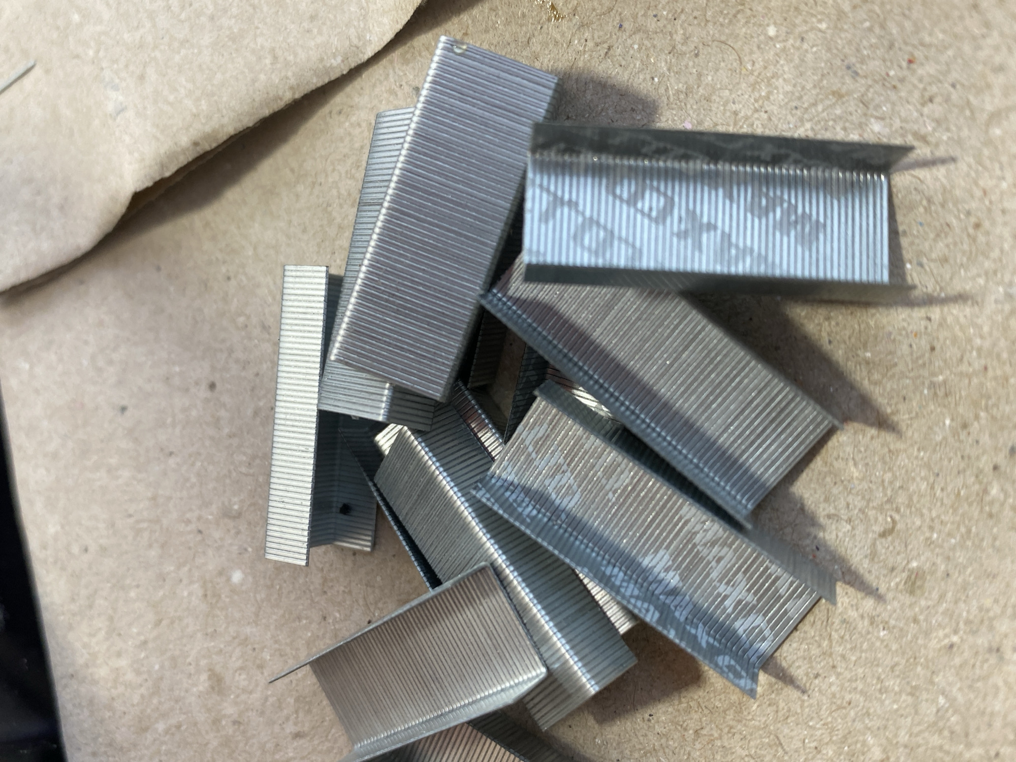 A pile of silver staples is scattered on a light brown, textured surface that appears to be cardboard or wood. The staples are rectangular and have parallel ridges on their surfaces. Some staples are partially obscured by others, creating a sense of depth and disarray. The lighting is natural, casting subtle shadows on the surface. There are no people or animals visible in the image. The location is not specified in the image or its metadata, and therefore cannot be determined.