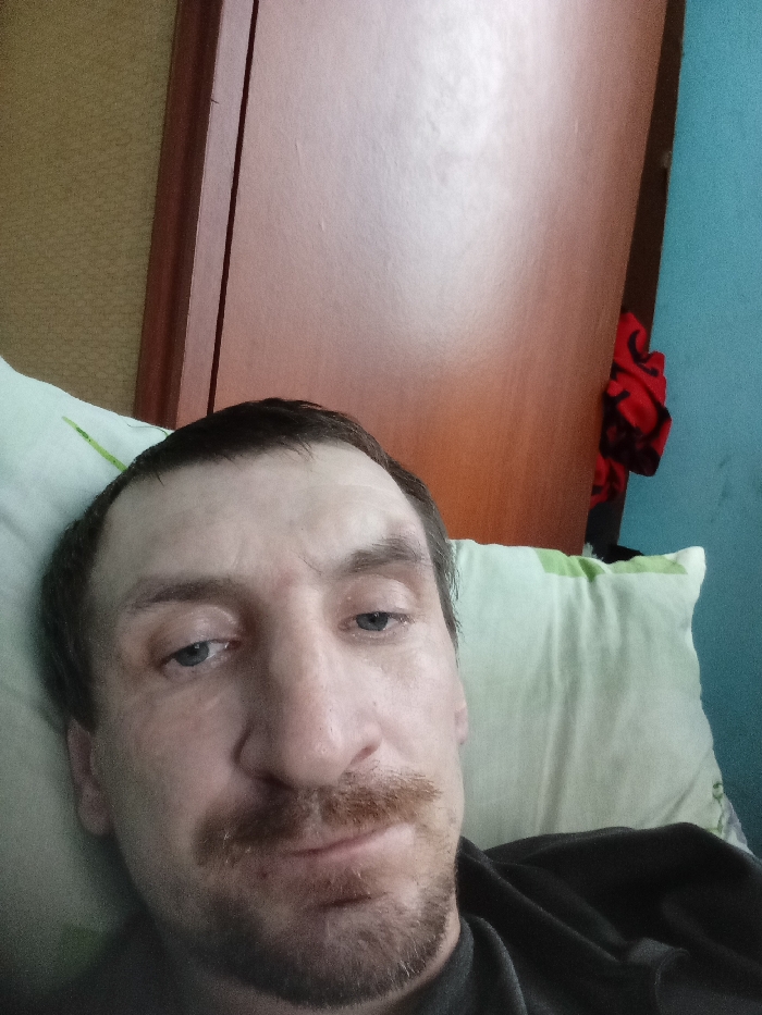 A male individual is depicted in a close-up, upward-angled shot, presumably a selfie, taken indoors within a residential setting in Yarun, Ukraine. The subject, a man with short, dark brown hair, a reddish-brown mustache, and a goatee, is lying down with his head resting on a light green pillow featuring a faint, darker green pattern. His eyes, appearing blue or grey, are directed towards the camera. He is wearing a dark-colored, likely black or dark grey, shirt. The background features a textured light brown or beige wall on the left, and a smooth, light brown wooden door or wardrobe panel prominently in the upper center and right. To the far right, a section of a light blue wall is visible, with a crumpled piece of red fabric, possibly clothing or a blanket, partially obscured between the wooden panel and the blue wall.
