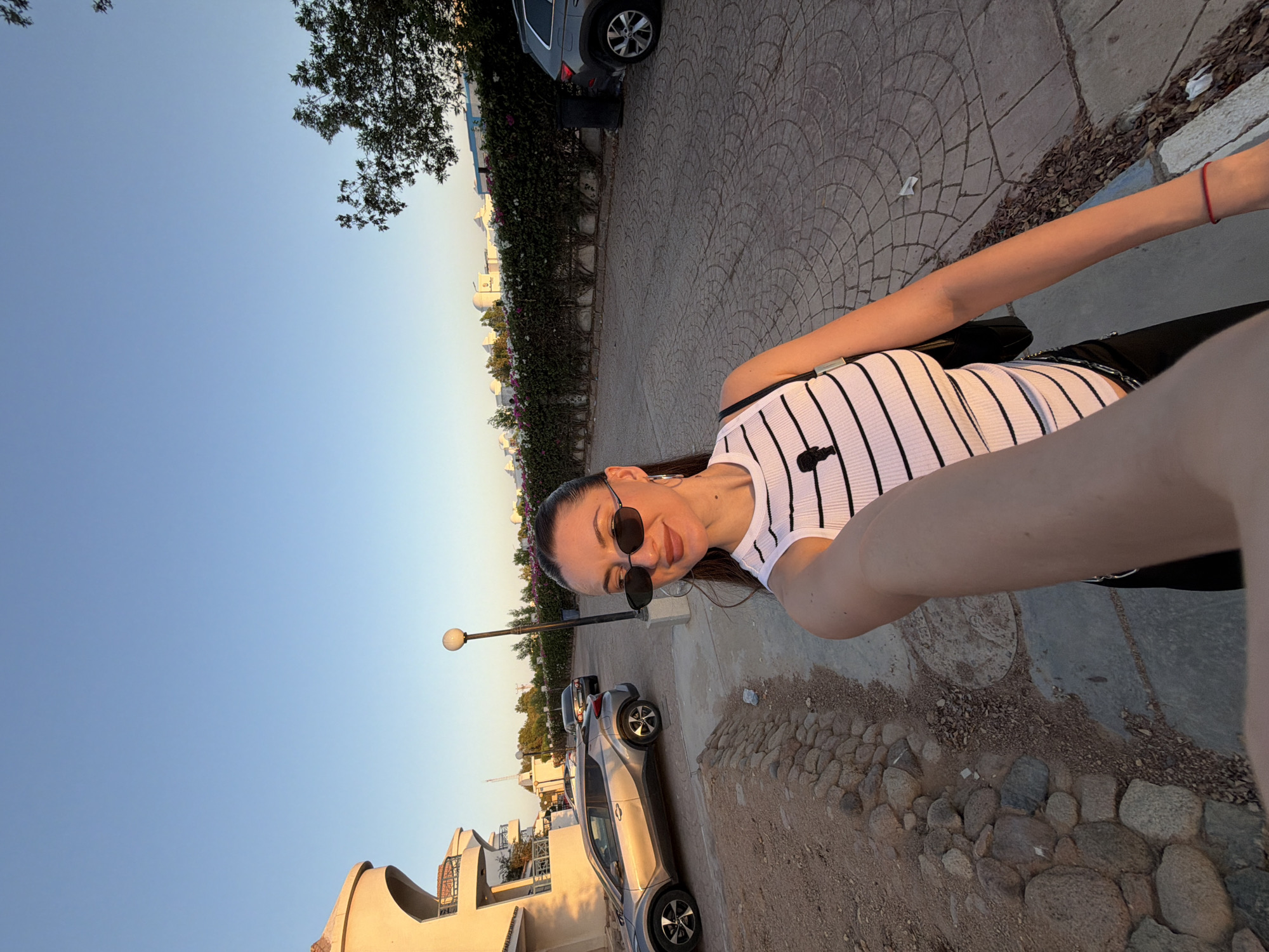 A full-shot, selfie-style photograph captures a woman standing outdoors in Sharm El Sheikh, Egypt, during what appears to be the golden hour. The woman, positioned in the foreground, is looking towards the camera with a slight smile. She has dark hair pulled back, is wearing black oval sunglasses, and large hoop earrings. Her attire consists of a white ribbed tank top with black horizontal stripes and a black design element on the chest, along with black pants. She is holding her phone out to take the picture with her right arm, and her left arm is by her side. A black purse with a chain strap is slung over her left shoulder.
The environment suggests a resort or residential area. To the left of the woman, a modern, cream-colored building with rounded balconies and arched windows is visible. A silver compact SUV is parked in front of the building. Further back, a darker sedan is parked, and beyond that, more vegetation and buildings can be seen. The ground in front of the woman is paved with textured, circular stone patterns. A narrow, rocky border separates the paved area from a dirt and gravel path. A lamppost stands near the center of the frame, behind the woman. A dense hedge with pink and white flowers runs along the right side of the scene, behind which a dark gray SUV is parked. The sky is a clear, pale blue, indicating a pleasant daytime or early evening setting. The overall scene is brightly lit by natural sunlight.