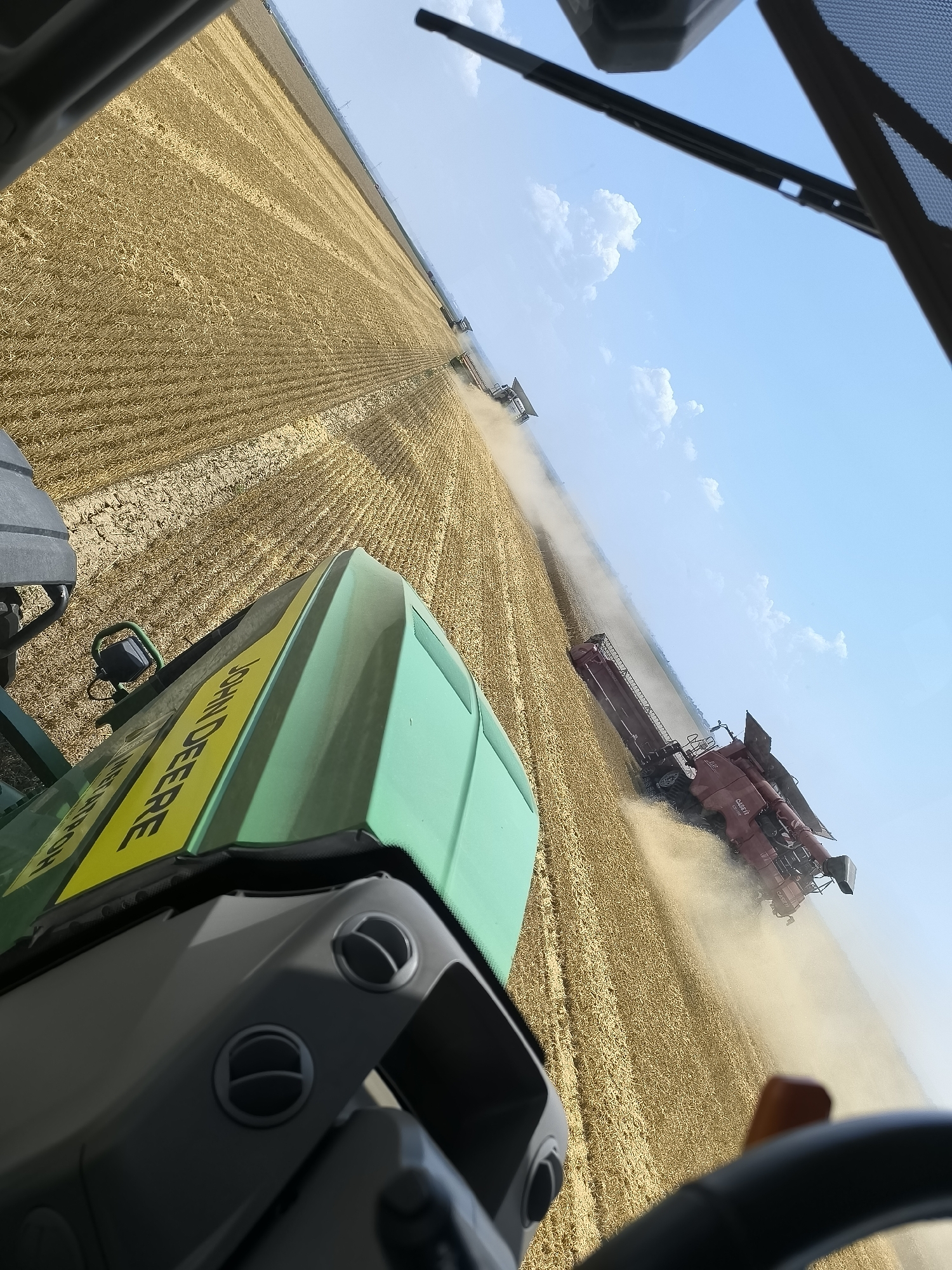 This media file captures a scene of agricultural harvesting in a vast golden field, likely wheat, under a bright blue sky with scattered white clouds. The perspective is from inside the cab of a large green tractor, identified by the "JOHN DEERE" text visible on its hood. The tractor's interior, including the dashboard and air vents, is in the foreground.
Several red combine harvesters are actively engaged in the field, working in unison to cut and gather the crop. One prominent combine, a "CASE IH" model, is clearly visible in the mid-ground, kicking up a significant amount of dust as it operates. This suggests dry conditions, typical for a harvest. The rows of harvested crops are clearly defined, indicating the progress of the machinery.
The scene depicts a large-scale farming operation, likely taking place during the daytime due to the bright sunlight and clear sky. The location context provided is Maior Uzunovo, Bulgaria. The overall impression is one of productive agricultural activity, showcasing modern machinery at work in a rural setting. There are no people visible in the media file.