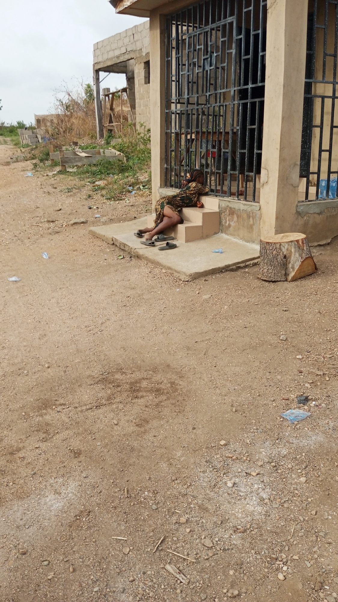 A woman is lying on the steps of a small building in Zango-ndaji, Nigeria. The building has a barred window and a concrete foundation. The woman is wearing a patterned dress and has her head resting on the steps. Her sandals are lying on the ground next to her. The ground is dirt and gravel. A tree stump sits next to the steps. Another partially constructed building is in the background. Plastic waste is scattered on the ground.