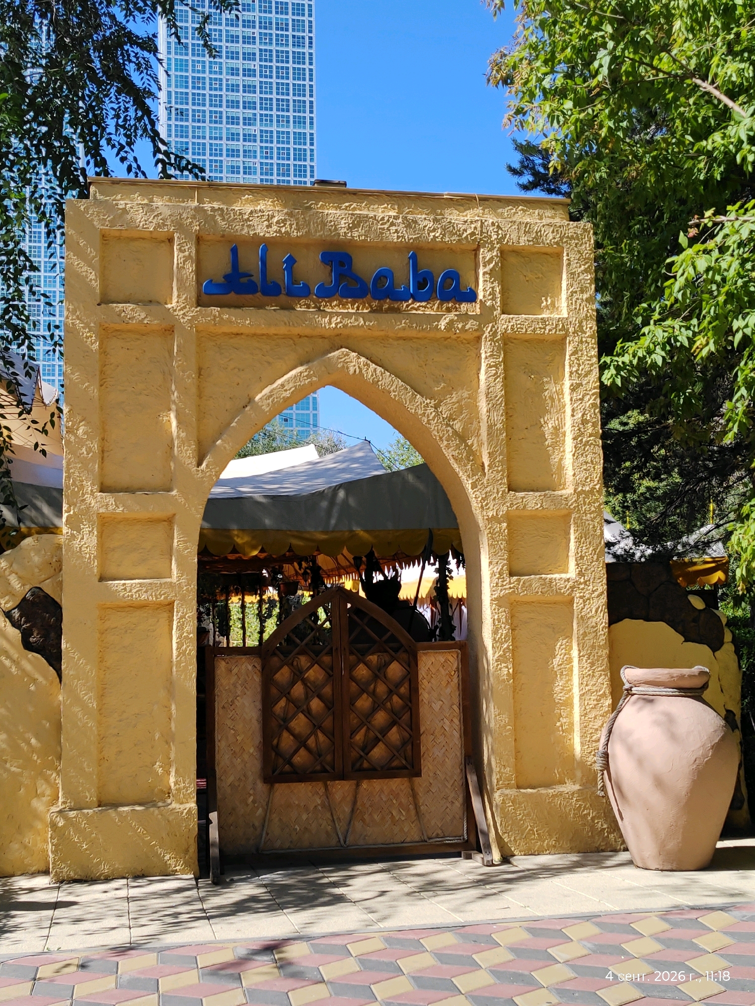The image shows an entrance archway decorated in a desert-themed style. The archway is painted a mustard yellow color and has a rough, textured finish. Above the arch, the words "Ali Baba" are written in blue Arabic script. Through the archway, tents and what appears to be a seating area can be seen. To the right of the arch is a large, clay amphora. In the background, a modern skyscraper with a grid-like facade is visible, contrasting with the traditional entrance. Lush green trees frame the scene. The ground in front of the arch is paved with colorful interlocking tiles. A timestamp in the bottom right corner reads "4 сент. 2026 г., 11:18", suggesting the photo was taken on September 4, 2026, at 11:18 AM. The overall impression is an outdoor establishment that combines a unique architectural style with a modern urban backdrop, possibly a restaurant or entertainment venue in Astana, Kazakhstan.