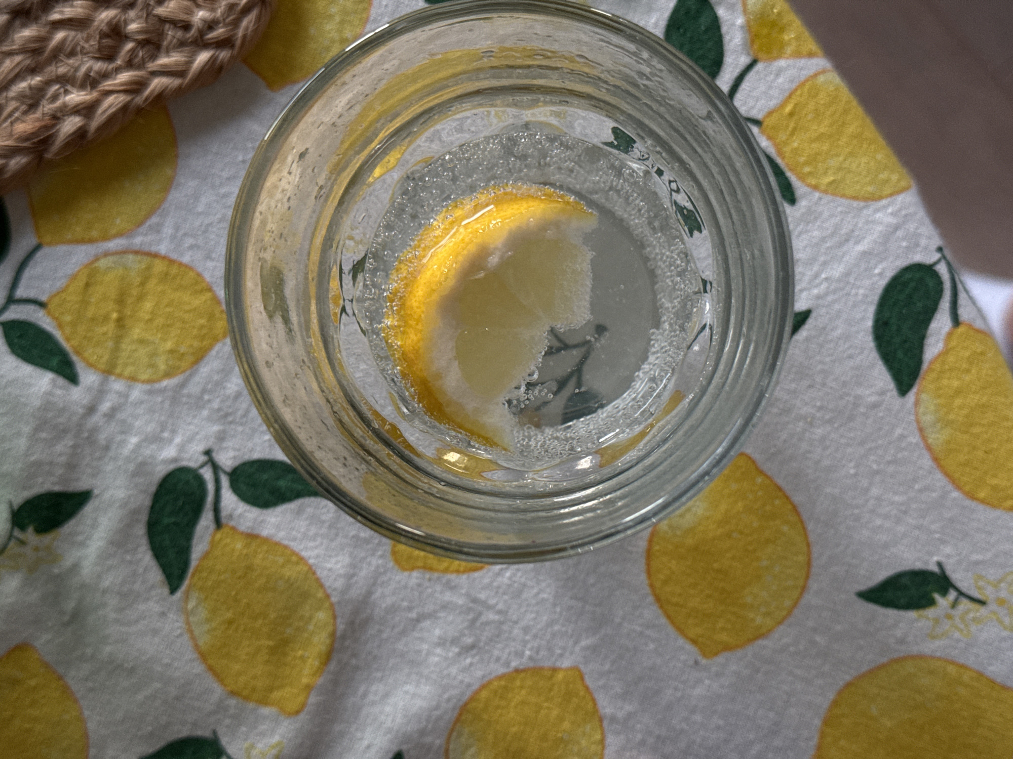 An overhead shot captures a clear glass filled with sparkling water and a single lemon slice, positioned on a light-colored tablecloth. The tablecloth features a repeating pattern of stylized yellow lemons with green leaves and small white blossoms.
Inside the glass, numerous small bubbles cling to the sides and the submerged lemon slice, indicating carbonation. The lemon slice is bright yellow with some white pulp visible. In the upper left corner of the frame, a portion of a woven, textured item, possibly a coaster or placemat made of straw or jute, is visible. A small section of a person's hand or arm can be seen in the upper right, casting a faint shadow. The scene suggests a refreshing drink, likely indoors in Vozdovac Urban Municipality, Serbia. No text is visible in the image.