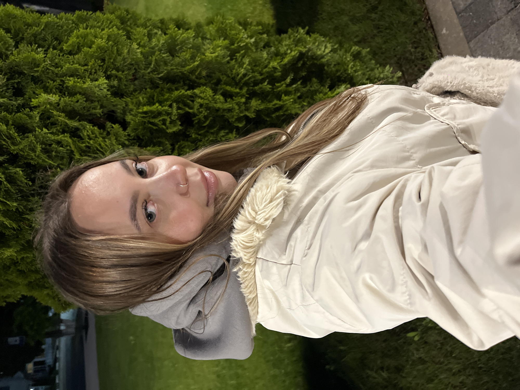 A young woman poses for a selfie outdoors at night. She is wearing a light beige jacket with a fuzzy collar and a gray hooded sweatshirt. The background includes a dense green bush and a dark area that appears to be grass and possibly a street with buildings visible in the distance. While the location cannot be confirmed solely by the image, the request mentions Novopokrovskaya, Russia.