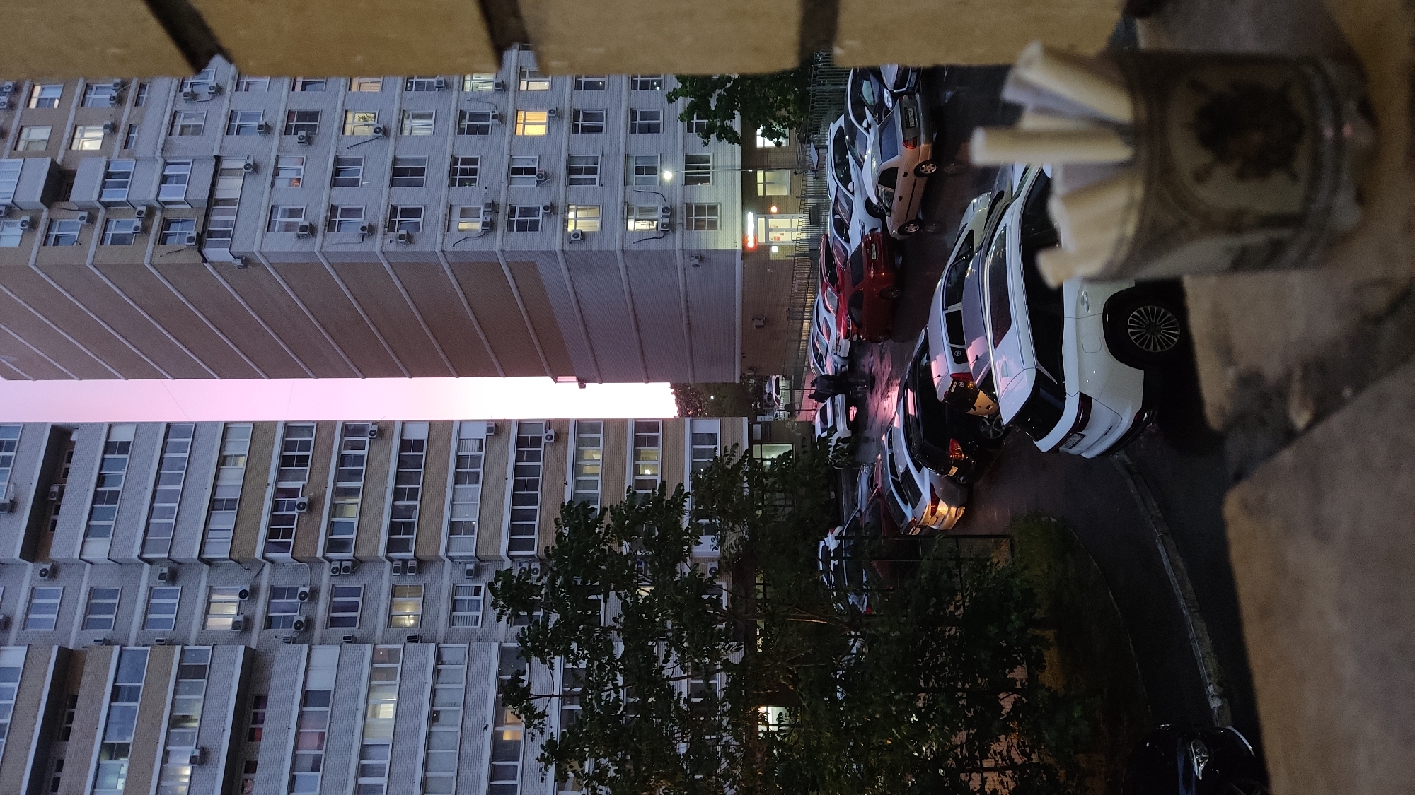 The image depicts a view from an elevated position, likely a balcony or high window, overlooking a parking lot and apartment buildings. The photograph appears to be taken at dusk or early evening, with the sky exhibiting a pinkish hue between the buildings. Two large apartment buildings dominate the left side of the frame. They are constructed from a light-colored brick or concrete and feature rows of windows with numerous air conditioning units attached. The buildings seem to have 10 or more floors. The parking lot is located to the right of the buildings and contains several parked cars. Most of the visible vehicles are white. There are also some trees in the foreground, partially obscuring the view of the parking lot and buildings. In the very foreground, there is a wall with a container of cigarettes on top of it. The location appears to be in Krasnodar, Russia.