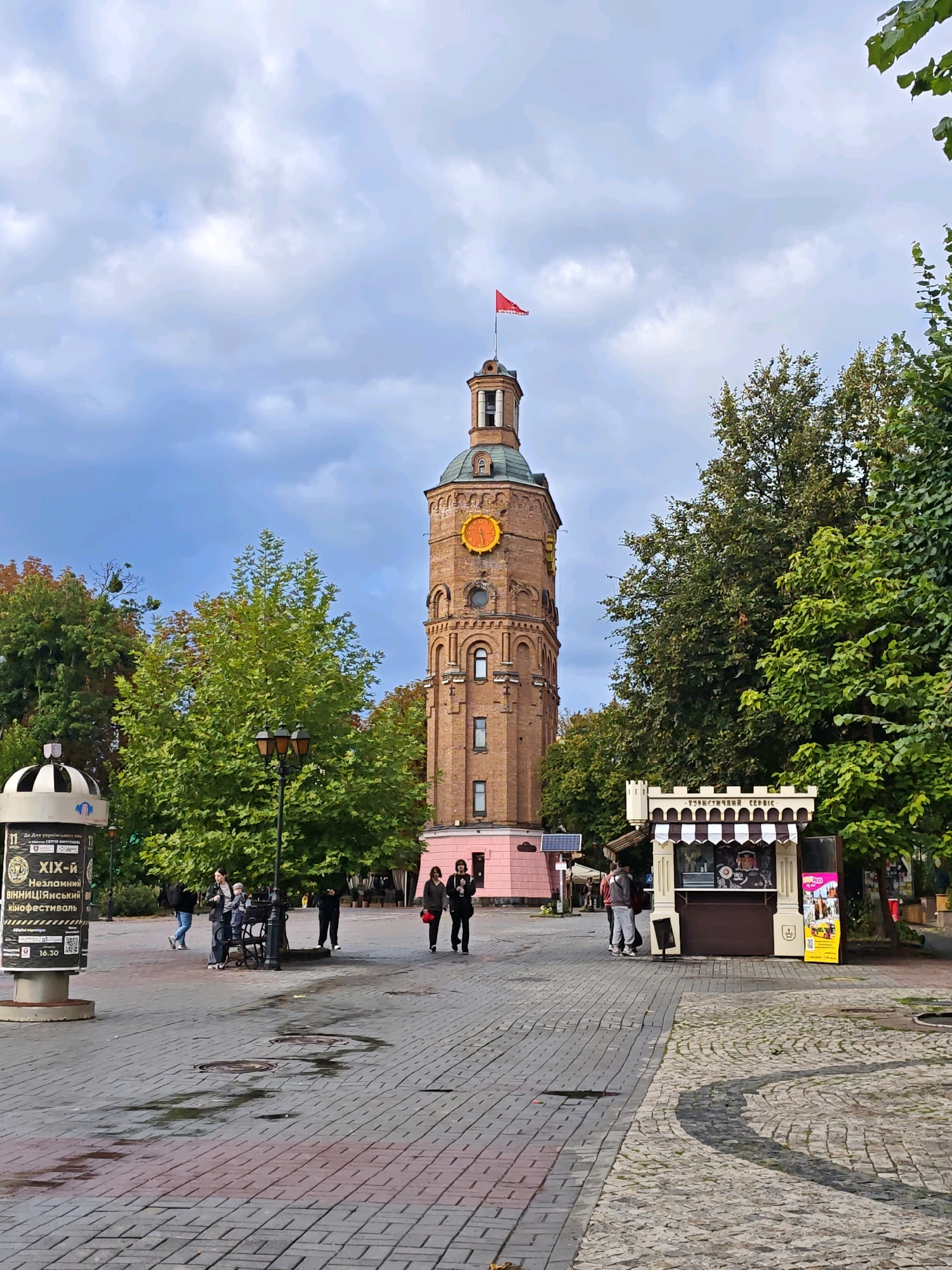The image captures the iconic Vinnytsia Water Tower, a prominent landmark in Vinnytsia, Ukraine. The tower, constructed from reddish-brown bricks, stands tall against a cloudy sky, topped with a red flag. A large, orange clock face is prominently displayed on its facade. At the base of the tower, a small pink building is visible.
The foreground features a paved pedestrian area with patterned brickwork, some sections appearing wet from recent rain. A lamppost with three lights stands on the left side of the image. To the right of the tower, a small, ornate kiosk with a striped awning is set up, appearing to sell food or drinks. Several people are present in the scene, walking and standing in the plaza. A cylindrical information stand with posters is also situated on the left. Lush green trees surround the plaza, adding to the urban park-like atmosphere. The overall impression is of a moderately busy public space on an overcast day.