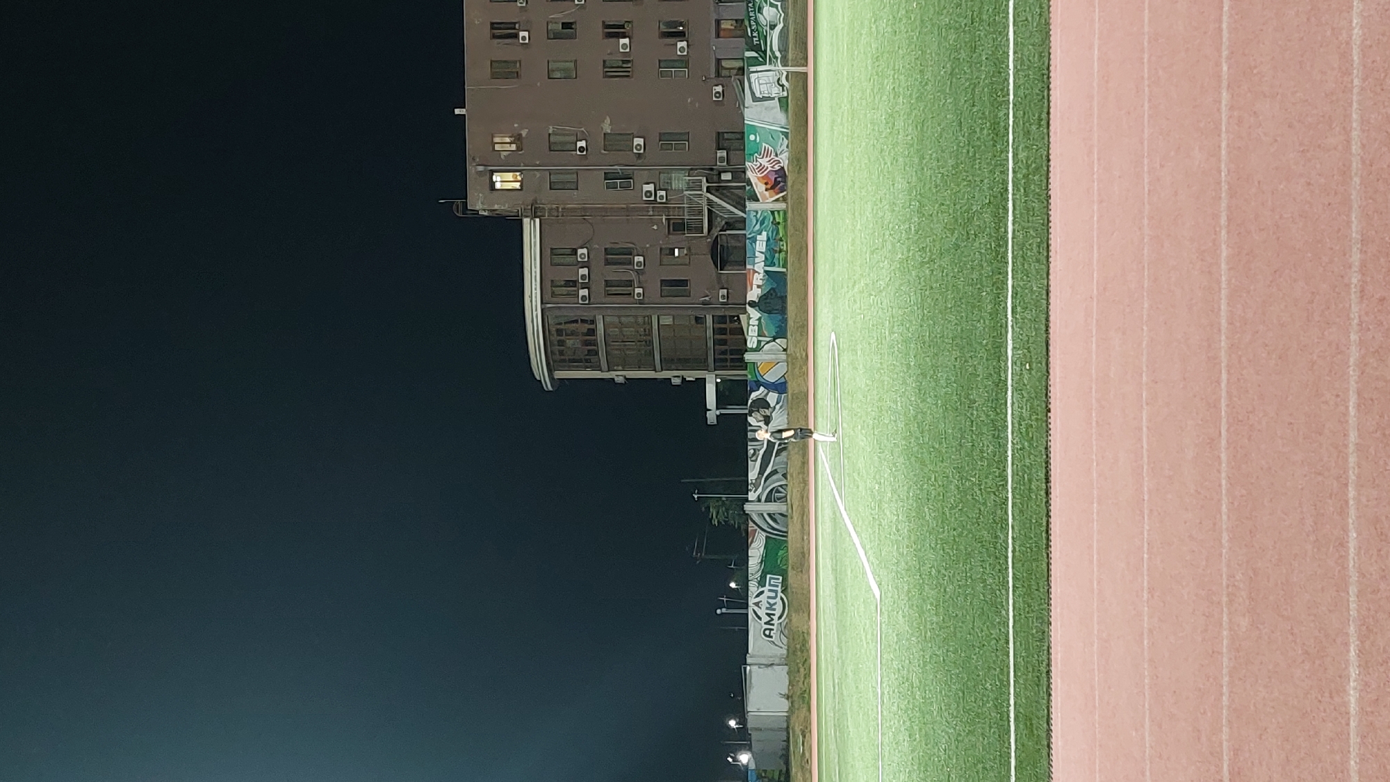 The image depicts an outdoor sports field at night, likely a soccer pitch, with a running track in the foreground. The field is illuminated, with white lines marking its boundaries. A lone figure, appearing to be a person in athletic attire, stands on the field. In the background, a multi-story building with numerous windows and air conditioning units is visible. The building has a distinctive rounded section. Behind the field and in front of the building, a mural or decorative wall runs along the perimeter, featuring various images and text, including the word "AMKUN." The sky above is dark. This scene is consistent with the sports complex located in Novocherkassk, Russia, a facility known for its athletic fields and surrounding infrastructure.