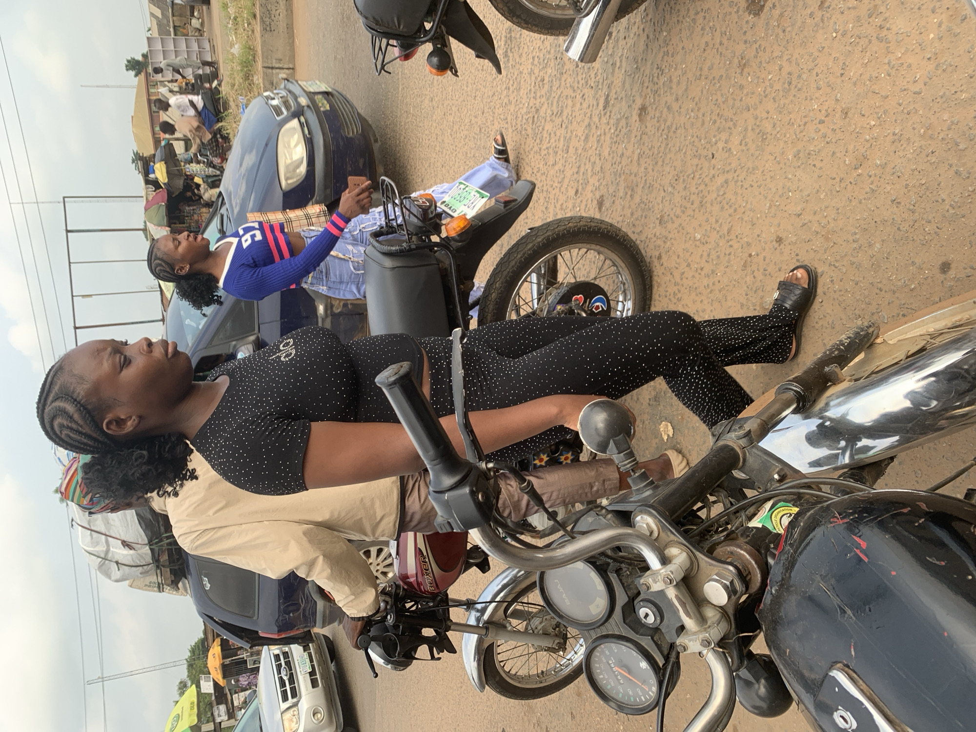 The image depicts a street scene in Shagamu, Nigeria, likely near a market area, with several motorcycles and vehicles. In the foreground, a black motorcycle is partially visible. To the right, another black motorcycle is seen from the rear. In the middle ground, a woman wearing a black, beaded jumpsuit and black sandals walks to the right. Behind her, another woman in a blue and pink crop top and light-colored jeans walks while looking to her left. A red motorcycle with a "Boxer" label is parked between them. Further in the background, a blue vehicle is parked, and a silver SUV is seen on the left. A man wearing a beige jacket and patterned headwrap stands between the vehicles. The ground is paved with rough asphalt and dirt. The sky is overcast. Market stalls with umbrellas are visible in the distance, suggesting a commercial or bustling urban environment.