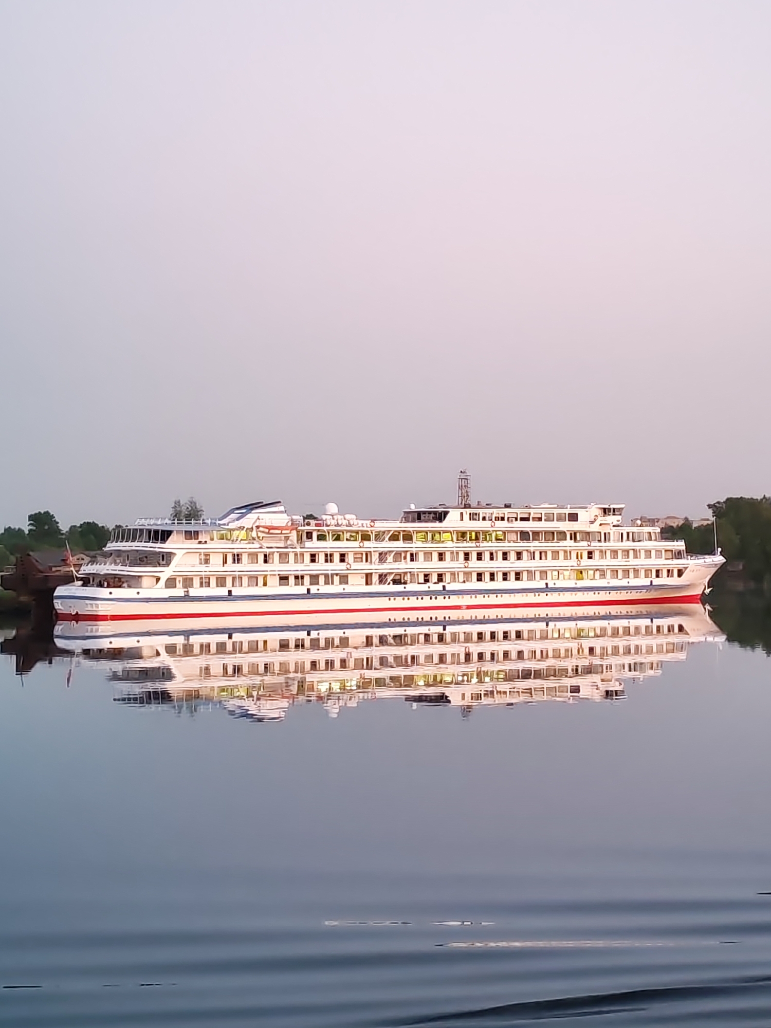 The image depicts a large, multi-decked river cruise ship docked along a calm body of water, likely the Volga River in Kimry, Russia. The ship is predominantly white with red trim, illuminated by interior lights, and reflected on the water's surface. A smaller vessel is docked alongside it. The sky is a pale grey, suggesting early morning or late evening light. Some trees are visible on the far bank.