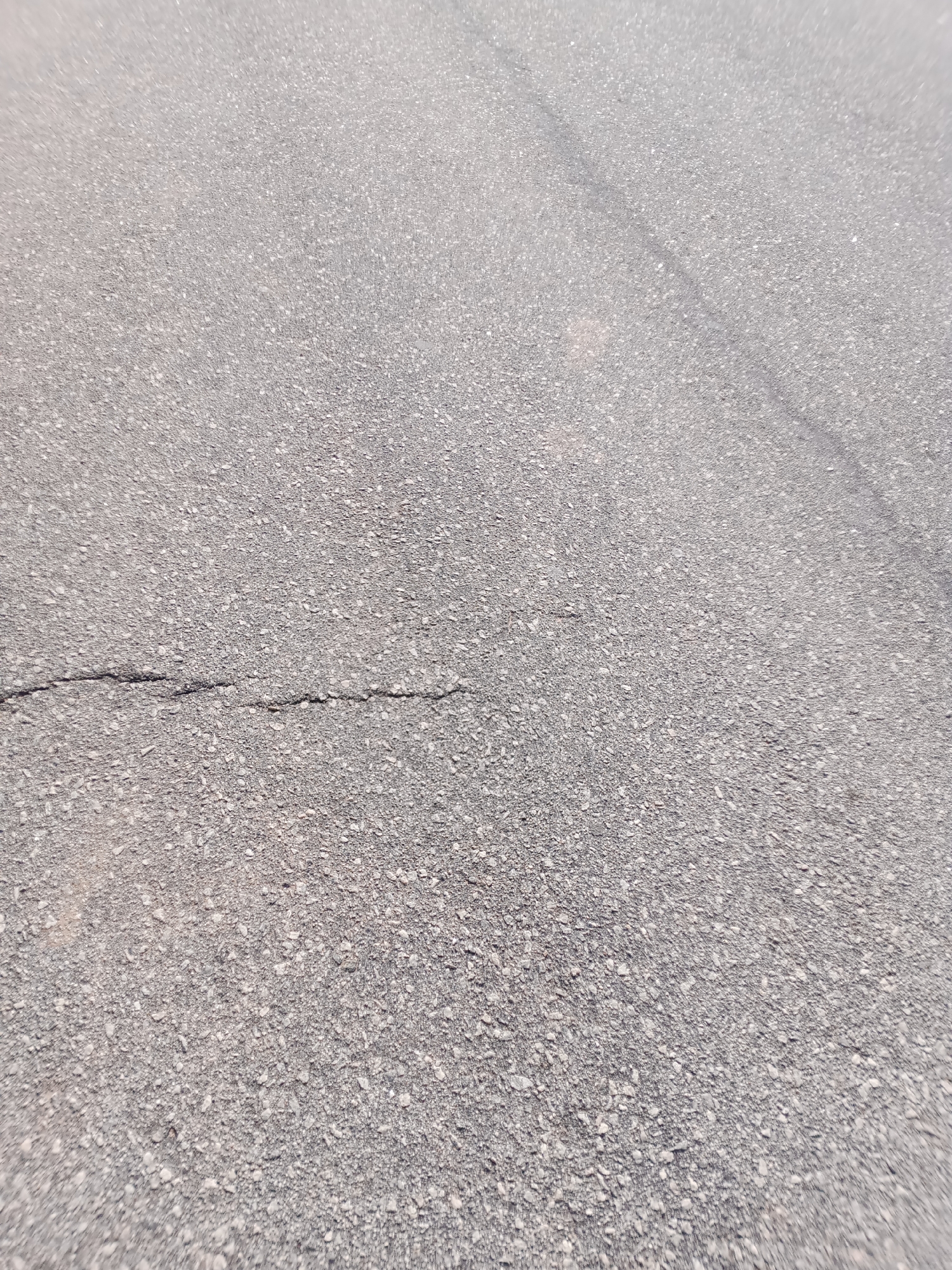 The image shows a close-up, high-angle view of an asphalt surface, likely a road or pavement, in Jalingo, Nigeria. The asphalt is grey and granular, with visible small stones and a subtle texture. A thin, dark, horizontal crack runs across the lower left portion of the image. In the upper right quadrant, a faint, diagonal line suggests a seam or a different section of the pavement. The overall impression is that of an outdoor, urban or semi-urban environment, characteristic of Jalingo. No people, vehicles, or specific landmarks are discernible, with the focus being solely on the ground surface.