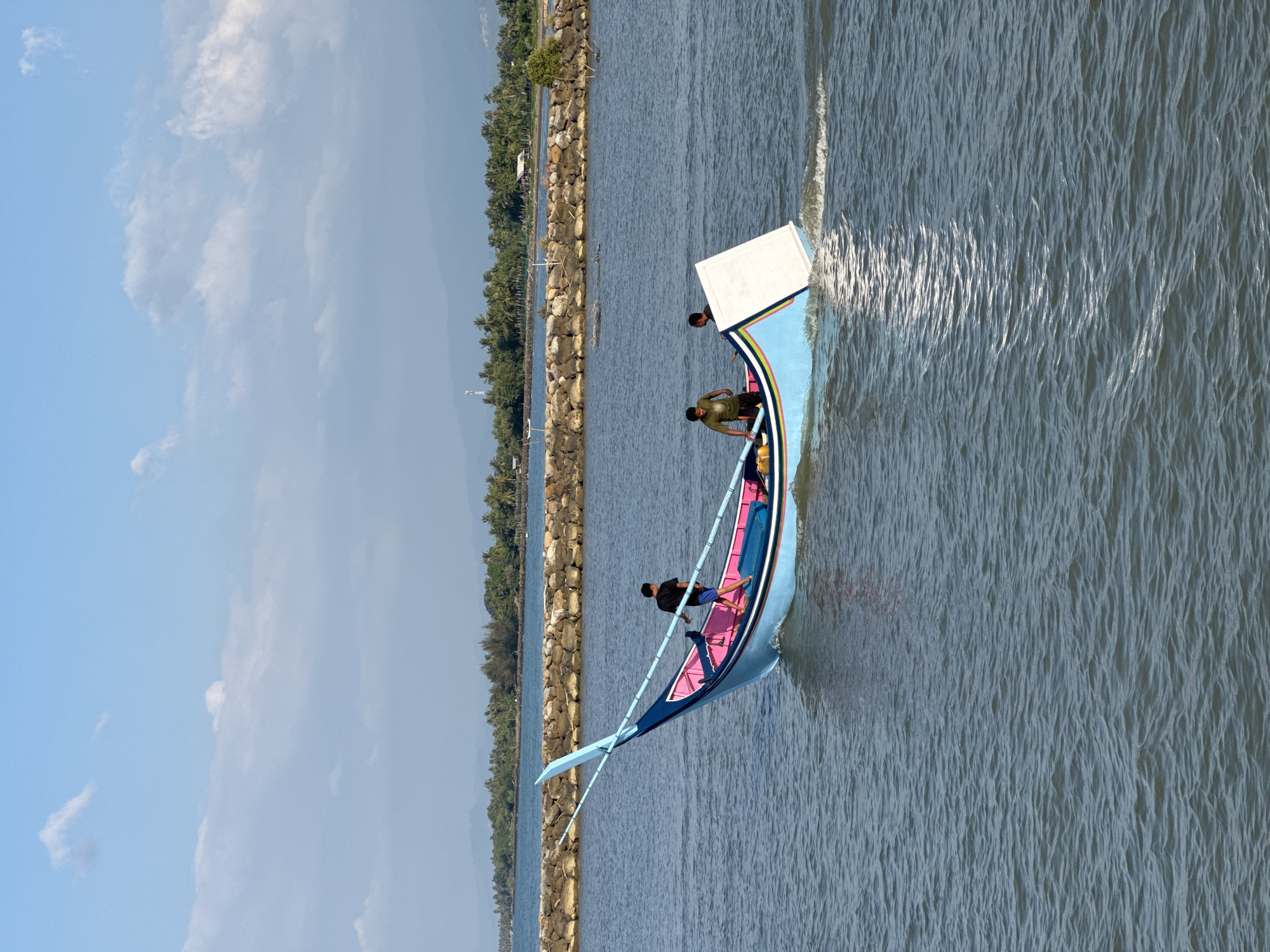 A light blue and pink boat with three individuals on board moves across a body of water. The boat has a long, slender design and appears to be made of wood. Two of the individuals are standing, while one is seated. The water is calm with some ripples. In the background, there is a stone embankment and a coastline with lush vegetation, including palm trees. The sky is clear with a few scattered clouds. The scene appears to be a harbor or coastal area, likely in Reudeuep, Indonesia, given the geographical context provided.