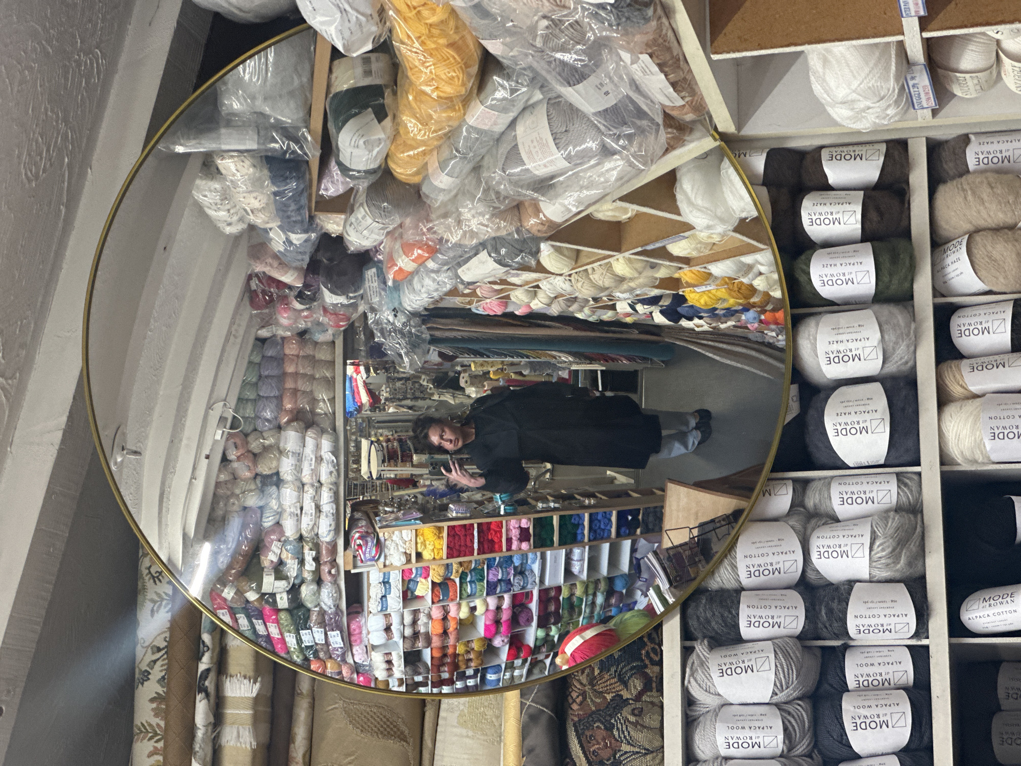 A woman is reflected in a gold-rimmed circular mirror surrounded by shelves of yarn. The mirror shows the interior of a yarn store. The shelves in the foreground contain rolls of yarn with labels. A woman in a black coat is taking a selfie in the reflection. The shelves reflected in the mirror display a wide range of yarn colors. Some fabric rolls are visible below the shelves in the mirror's reflection. The yarn labels indicate the store is in Sevenoaks, United Kingdom, selling "MODE at ROWAN" yarn.