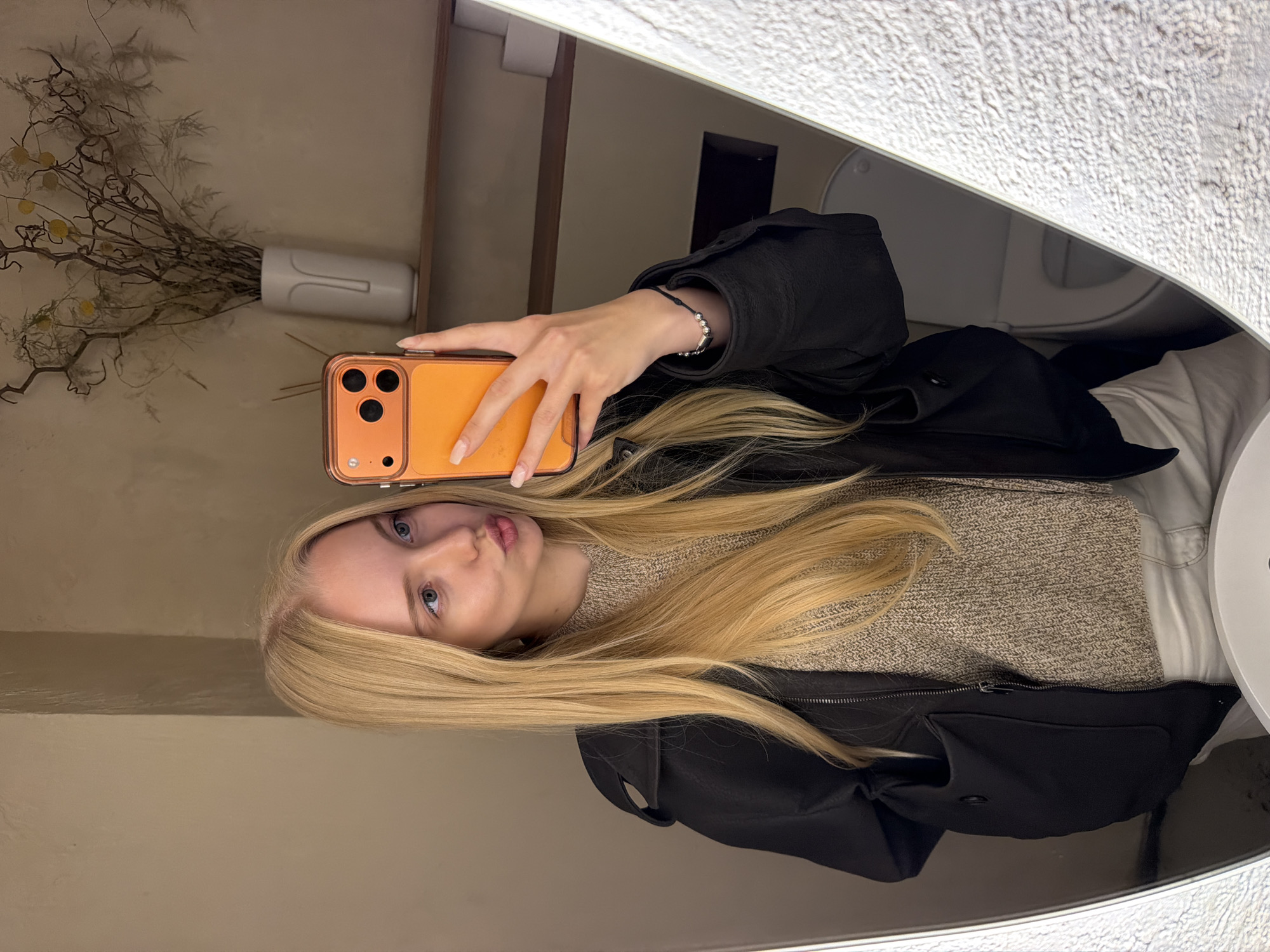 A young blonde woman, seen from the chest up, takes a mirror selfie indoors. She has light skin, light eyes, and is making a pouting expression. She wears a black jacket over a light brown knitted sweater and light-colored pants. Her right hand, holding a bright orange-cased smartphone, is adorned with a dark cord bracelet featuring silver beads. The background, reflected in the mirror, shows a smooth, light tan wall. On a wooden shelf, a white vase with a minimalist face design holds dried branches and foliage with small yellow accents. Below the shelf, a toilet is partially visible in the lower right, along with two rolls of toilet paper on the shelf, indicating a restroom setting. The mirror itself has a textured, light-colored frame on the right side. The location is Unknown, Unknown.