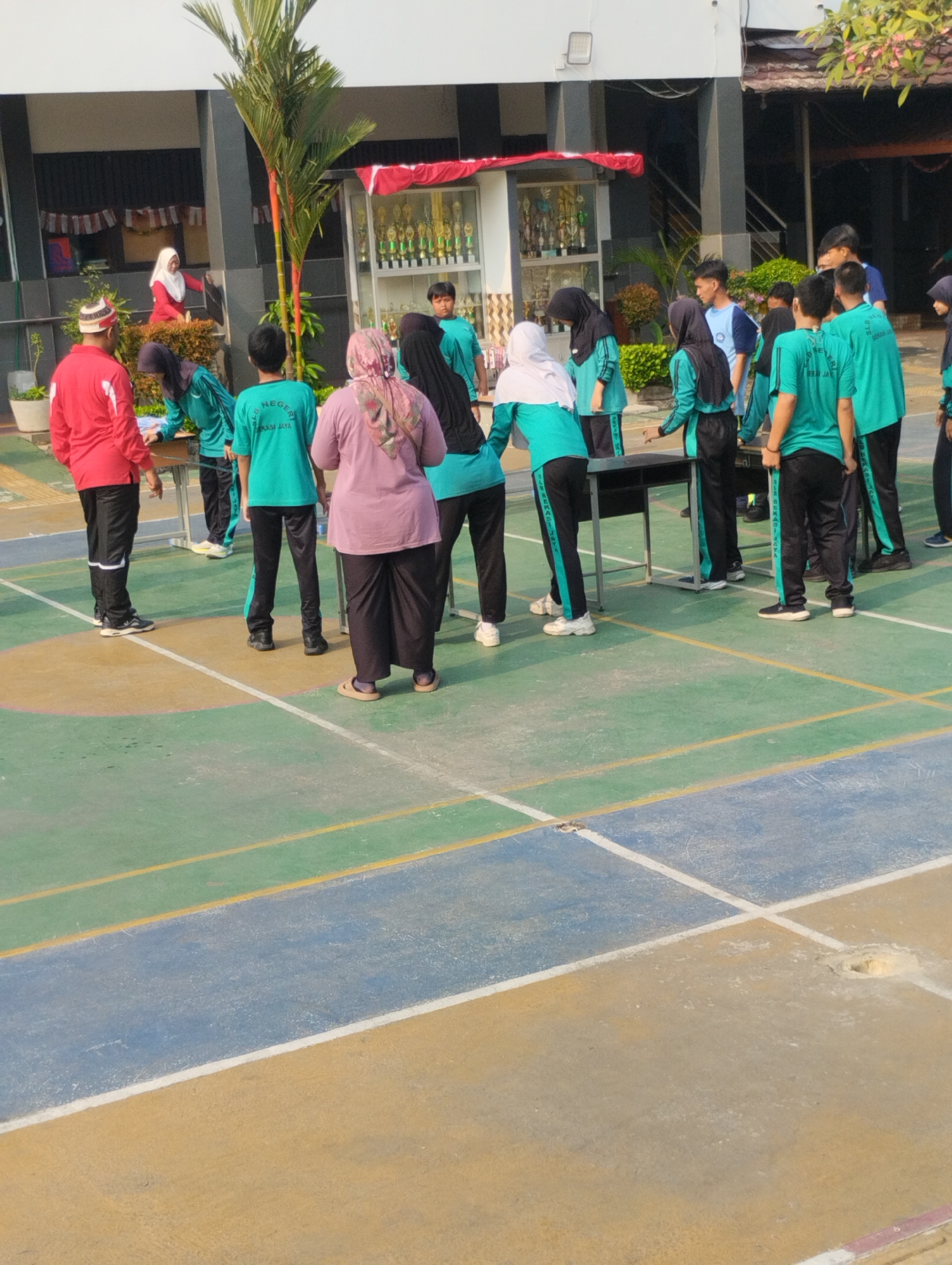 In front of a school building in Bekasi, Indonesia, a group of students and adults are gathered on a multi-colored court, possibly for an event or activity. Several individuals are wearing teal colored uniforms with black pants, while others are in casual clothing. One person in a red shirt and black pants stands near a table. A woman in a pink headscarf and black pants is also visible. In the background, a trophy display case is visible inside the building. The court is marked with lines, suggesting it might be a sports facility. The sun is casting shadows, indicating it is likely daytime.