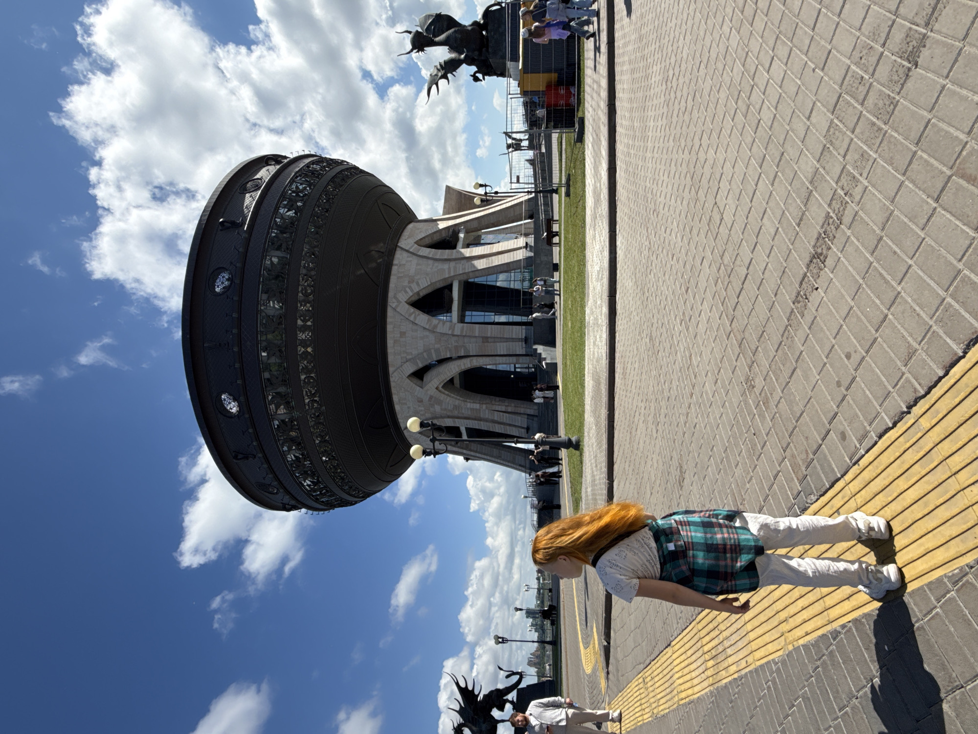 This outdoor image, presented rotated 90 degrees clockwise, depicts a distinctive architectural landmark under a bright, partly cloudy sky. The central feature is a large, dark, drum-shaped building adorned with intricate, decorative patterns and arched openings at its base. This structure is identifiable as the Kazan Family Center, often called "The Chasha," in Kazan, Russia.
In the foreground, a young person with long, reddish-blonde hair is seen in profile, walking on a light-colored paved pathway that features a yellow textured strip. They are dressed in a white short-sleeved top, a green and teal plaid vest or dress, light-colored pants, and white sneakers.
The area around the building includes green lawns and additional paved sections where other people are visible, some standing near the building's entrances and others strolling. To the left of the main structure, a dark, stylized sculpture of a mythical winged creature, consistent with the Zilant dragon symbol of Kazan, is present, with another similar sculpture partially visible further in the background.
The scene is brightly lit by the sun, indicating a clear, sunny day, likely mid-morning to afternoon, with scattered white cumulus clouds across a blue sky. The overall atmosphere suggests casual sightseeing or leisure activity around a public landmark. No discernible text is visible in the image.