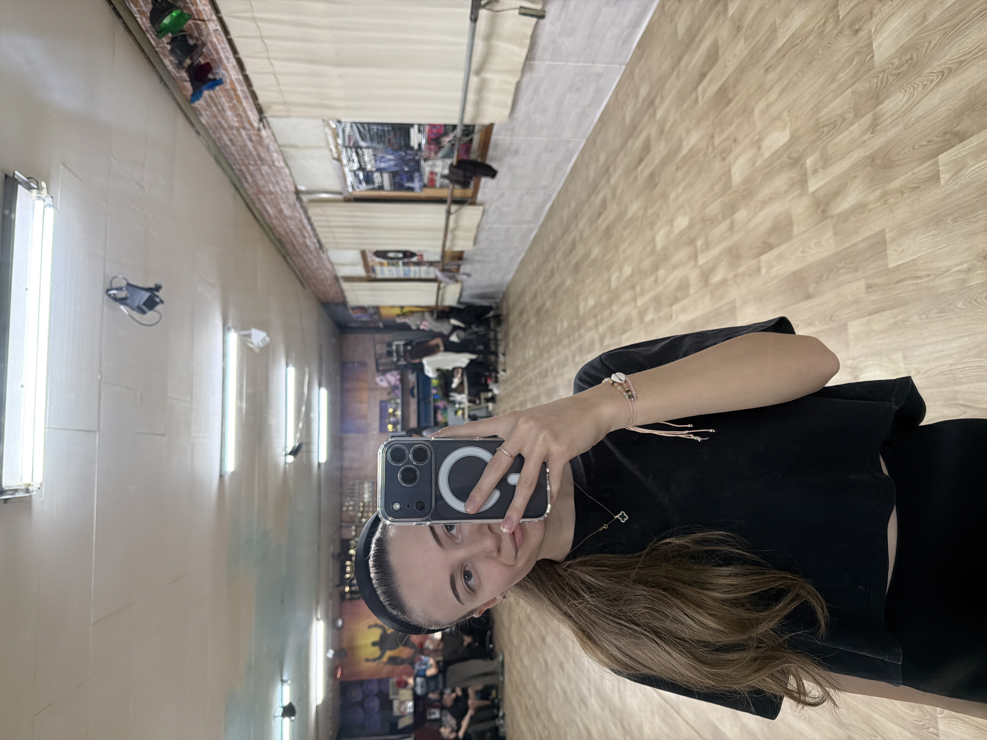 A young woman is taking a selfie in what appears to be a hair salon or beauty parlor, likely in Plovdiv, Bulgaria, based on environmental clues. She is holding a smartphone with a clear case, partially obscuring her face. She has fair skin, brown hair, and is wearing a black headband and a black top. The background reveals a long, narrow interior with multiple workstations and mirrors along the walls. Fluorescent lights are visible on the walls. There are other people and objects visible further down the hall, suggesting the space is occupied and in operation. The floor is a light-colored laminate. There's a mix of equipment and decorations typical of a beauty salon.