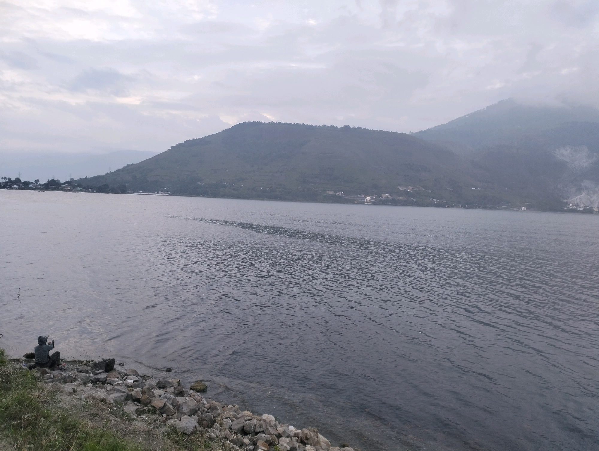 A person is seated on a rocky shoreline next to a large body of water. The person is wearing a dark hooded jacket and is facing towards the water, appearing to hold something in their hands. The shoreline is composed of various sized rocks and some sparse vegetation. The water is calm with a slight ripple. In the background, a large, vegetated mountain range stretches across the horizon under an overcast sky. The mountain slopes are dotted with some structures and trees, suggesting a settlement along the waterfront. The scene appears to be in a natural setting, possibly a lake or a bay, with a vast expanse of water and mountainous terrain. The location is identified as Parlondut, Indonesia.