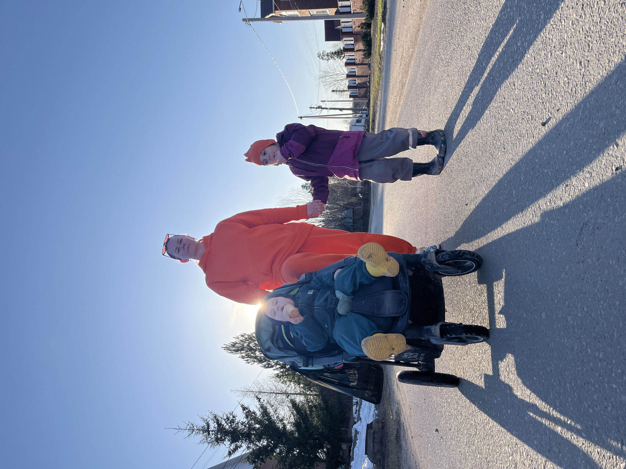 Three individuals, an adult and two children, are captured on a paved road in what appears to be Chaikovsky, Russia. The image is taken outdoors on a bright, sunny day, with a clear blue sky. The sun is positioned low in the sky, creating prominent, long shadows stretching across the road, suggesting either early morning or late afternoon.
In the foreground, an adult wearing an orange hooded sweatshirt with sunglasses perched on their head is pushing a dark-colored stroller. Their face is partially obscured by the bright sun behind them. Seated in the stroller is a young child dressed in a dark blue or green outfit and bright yellow shoes, holding what looks like a light-colored snack to their mouth. The stroller's frame shows some text, possibly "Mountain Buggy" and "all terrain."
Walking beside the adult and stroller is another child, wearing a purple and pink jacket, grey pants, dark boots, and an orange hat with small ear-like protrusions. They appear to be looking towards the camera.
The background features a residential street with buildings, utility poles, and scattered evergreen trees. Patches of snow are visible on the ground near the trees and buildings, indicating a cool season, likely late winter or early spring. The scene conveys a casual outdoor walk or excursion.