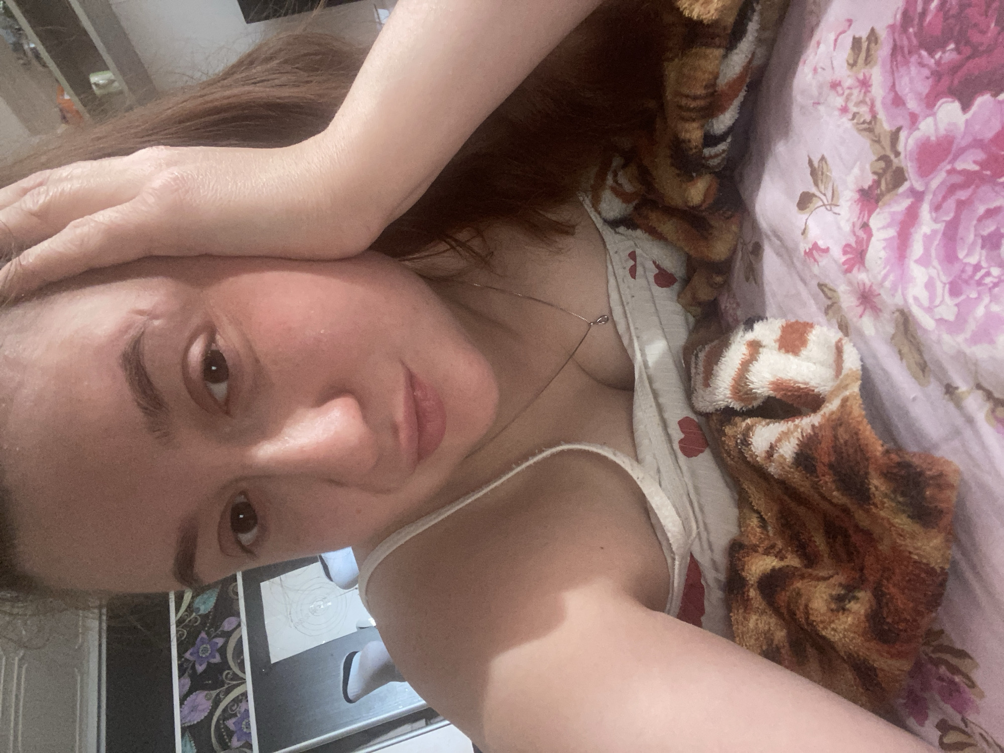 A selfie-style photograph shows a woman indoors. She is lying down, with her head resting on her hand. The background shows a floral patterned bedding and what appears to be part of an interior space with a kitchen counter. I am unable to verify a precise location within the city of Chaikovsky, Russia, without additional location information or metadata embedded in the image file.