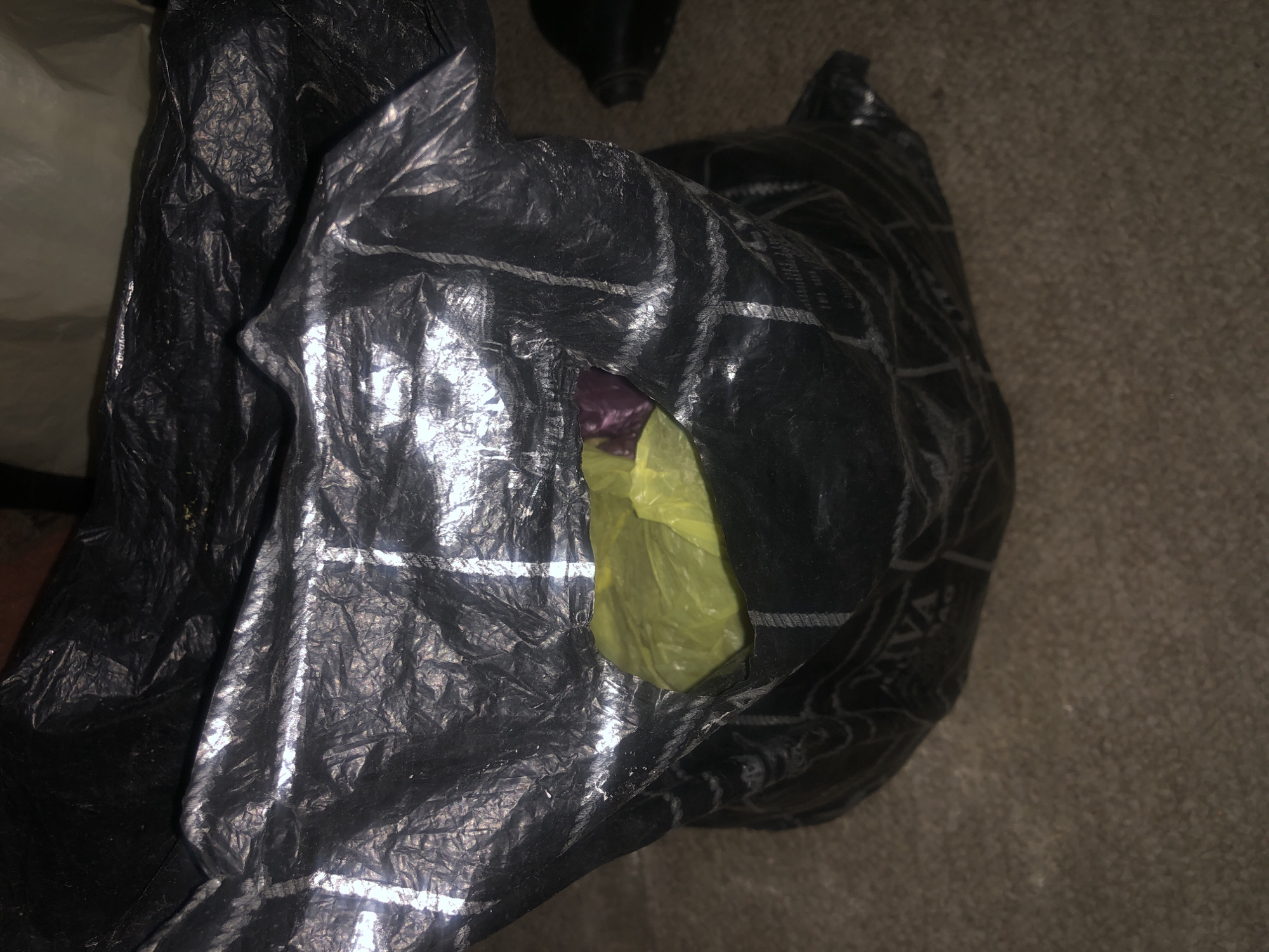 The image shows a close-up view of a black plastic bag on a brown carpeted floor. The bag is open and appears to be partially filled with other plastic bags, one of which is bright yellow and another is purple. The black bag has a reflective, crinkled surface with white lines patterned across it. The background is dark and out of focus, with a suggestion of other dark objects. No specific location in a city is discernible.