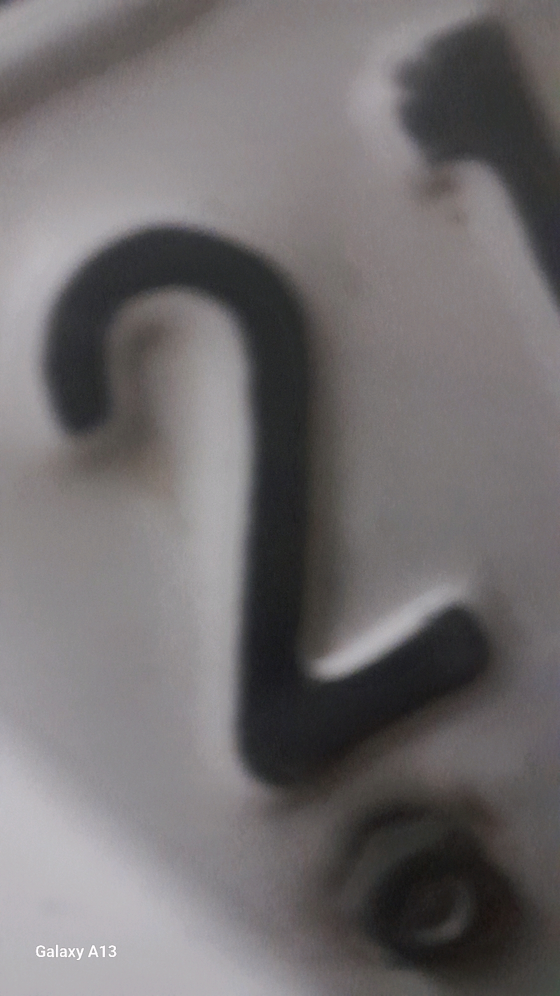 The image displays a close-up, out-of-focus view of a dark number "2" embossed on a light-colored surface. The surface appears to be metallic or hard plastic, with a slightly textured finish. The lighting is diffuse, casting soft shadows and highlighting the raised form of the numeral. The lower-left corner of the image shows the text "Galaxy A13," indicating the device used to capture the photograph. The background is indistinct, suggesting a shallow depth of field or a plain, uniform surface. The overall impression is of a detail captured from a larger object, possibly a sign, a plaque, or a piece of equipment. There are no people, actions, or interactions depicted, nor is there any identifiable urban environment.