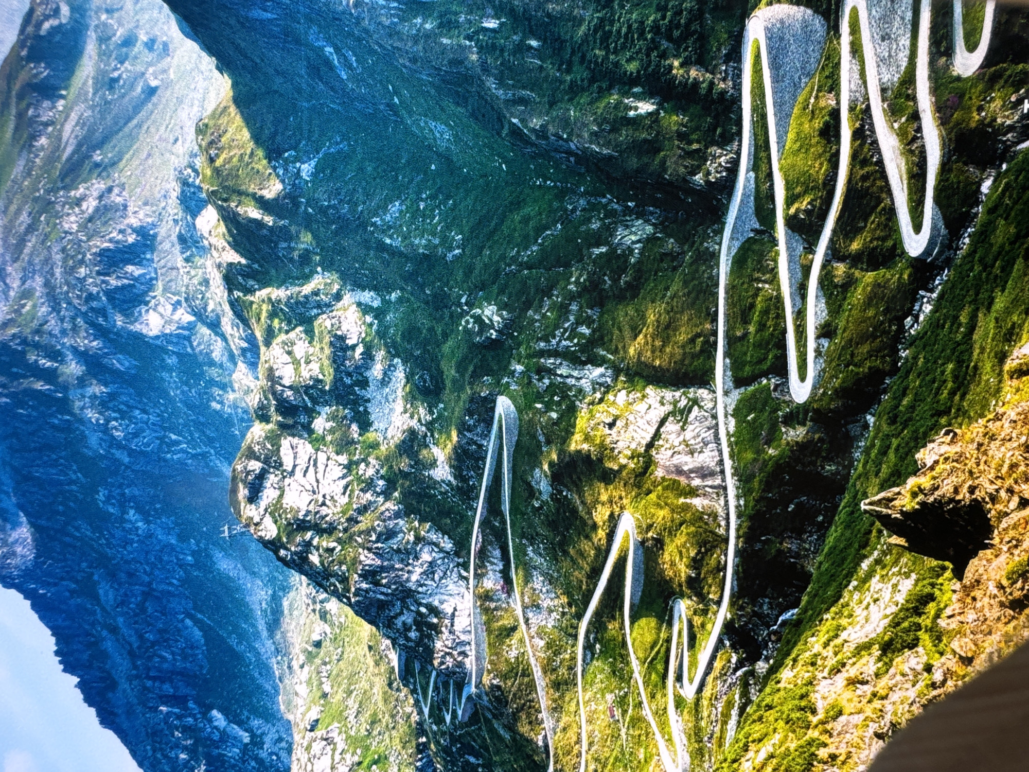 This image captures the dramatic switchbacks of the Susten Pass road in the Swiss Alps, near Schattdorf, Switzerland. The winding road, constructed with stone retaining walls, carves its way up the steep, verdant mountainside, illuminated by the sun. Patches of green grass and vegetation cover the rocky slopes, interspersed with stark white rock formations. In the distance, imposing, deep blue mountains rise, their peaks shrouded in shadow. The sky above is a bright, clear blue. There are no people or vehicles visible in the scene, emphasizing the grandeur and isolation of the natural landscape and the engineering feat of the road. A small, dark-colored structure, possibly a building, is visible nestled amongst the greenery along the road in the mid-ground.